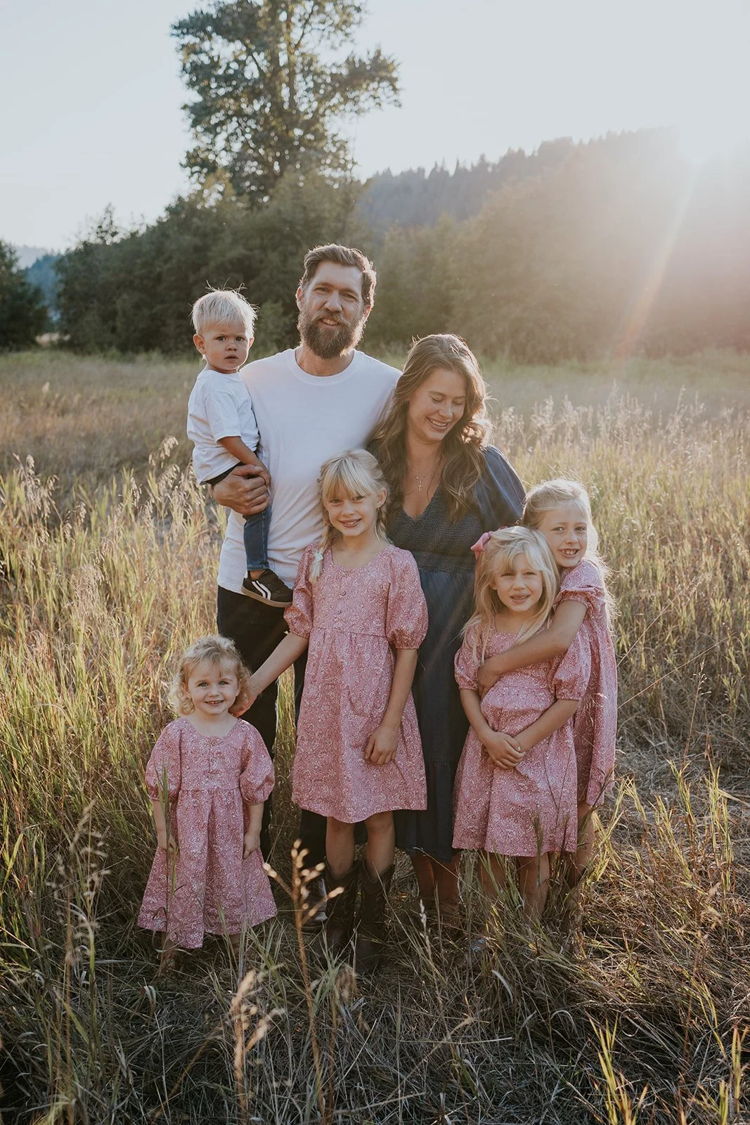 A family of seven, including adults and children, standing in a field with tall grass during sunset, smiling and embracing each other.