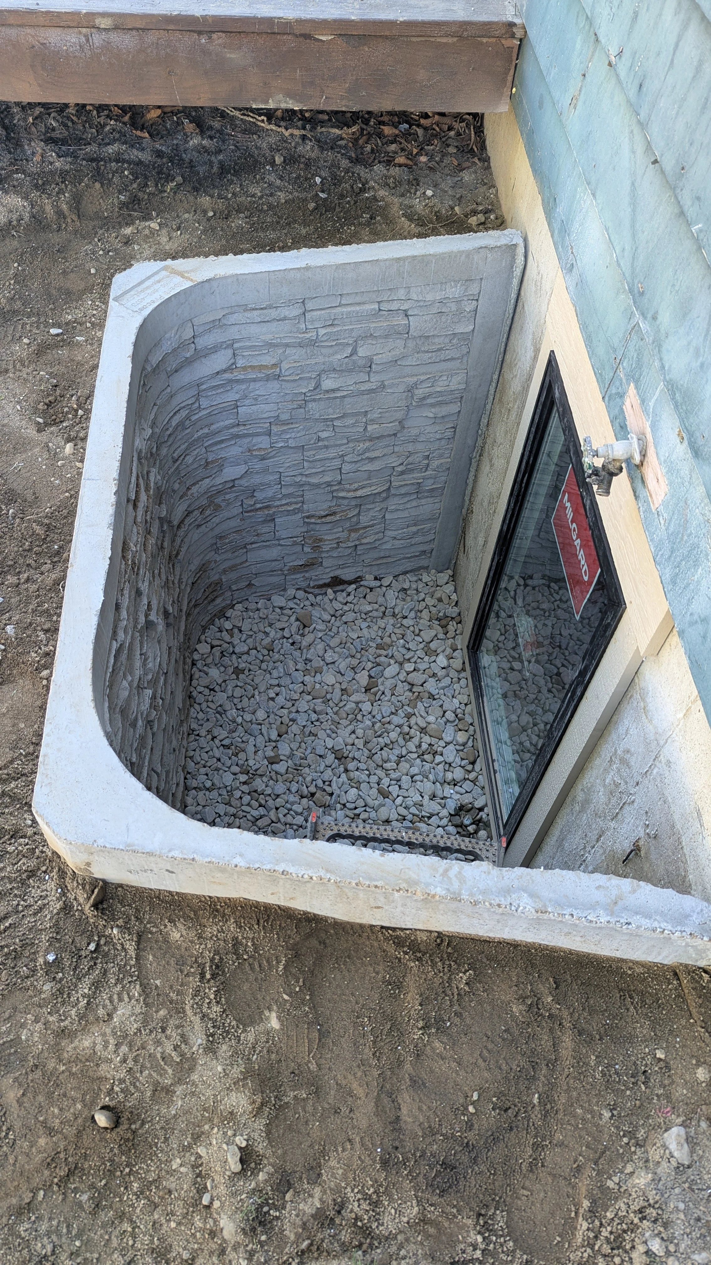 A basement window with a stone curved interior and gravel covering the ground inside the window well. The area around the window well has exposed dirt and a garden hose faucet on the outside wall.