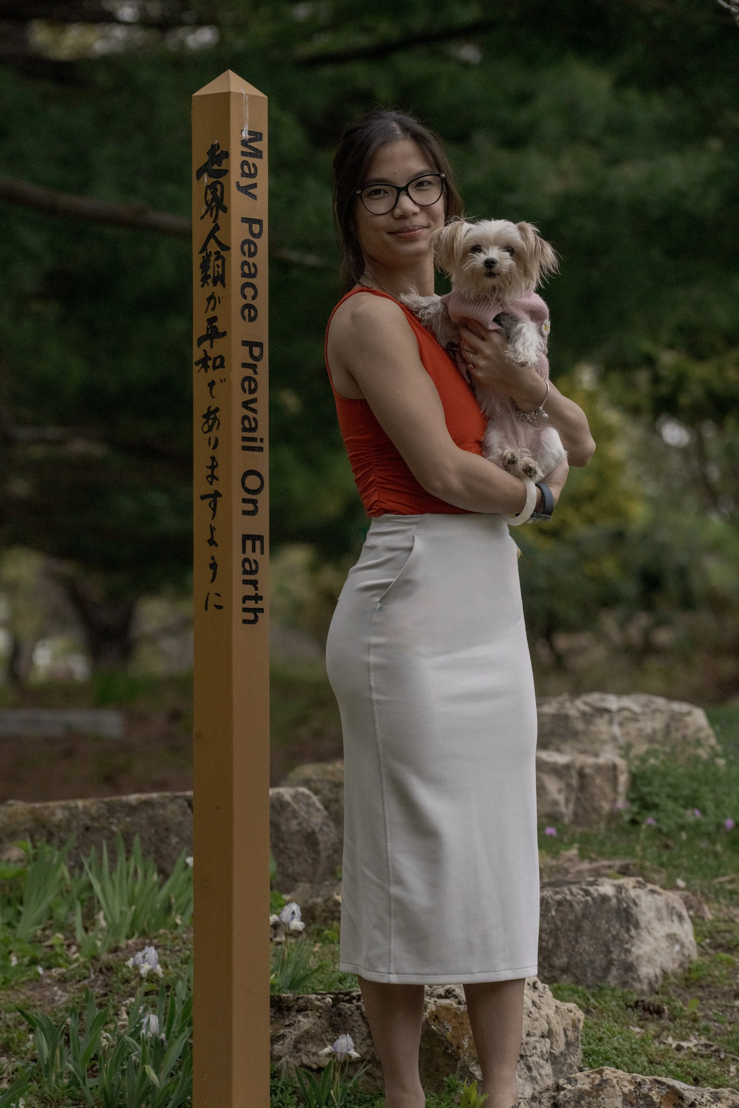 A woman wearing glasses and a sleeveless orange top holding a small dog wearing a pink sweater, standing next to a yellow peace on earth sign in a park with trees and rocks in the background.