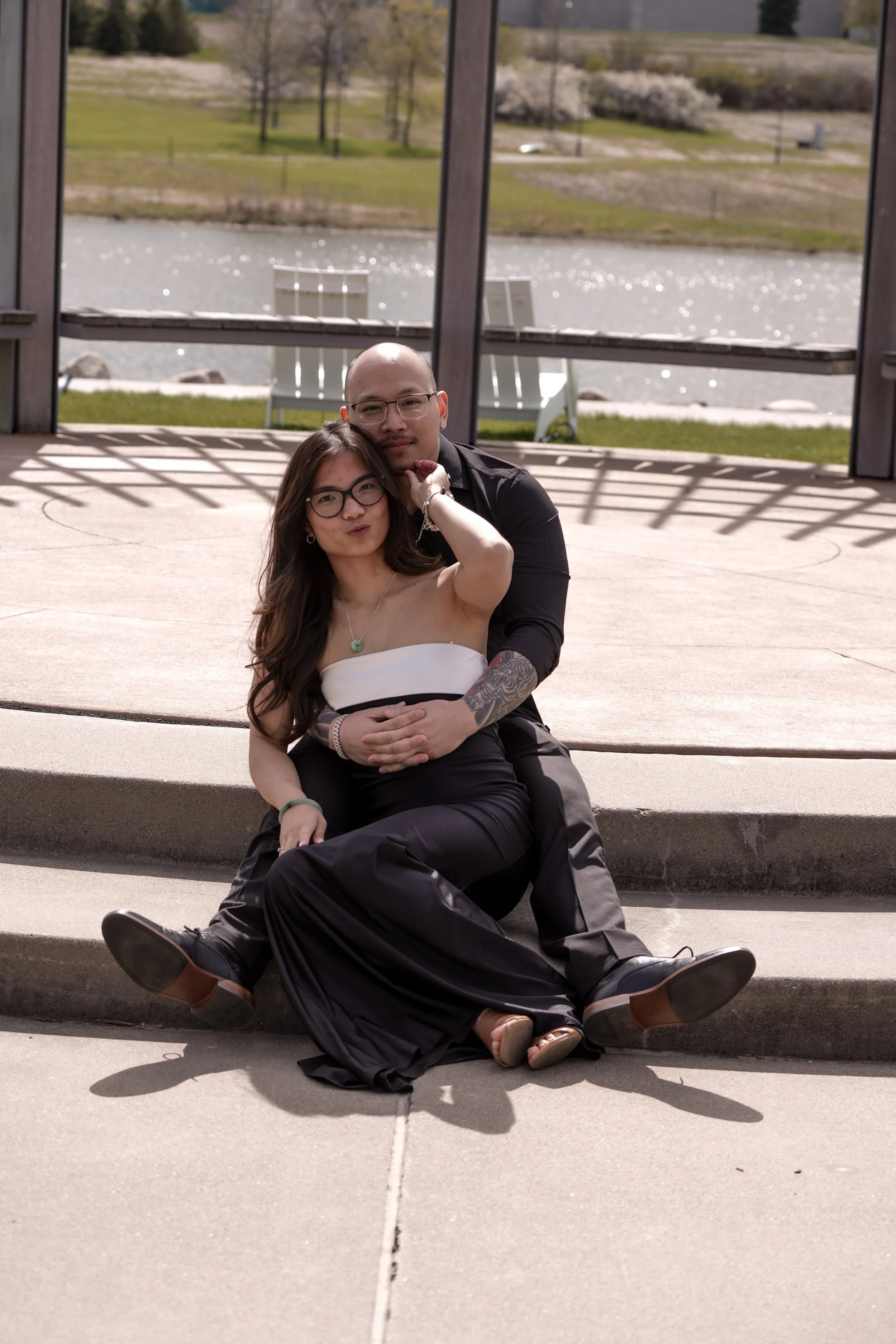 A couple sitting outside by a lake with trees and a grassy hillside in the background. The woman is wearing glasses, a white top, and black pants, with long dark hair. The man is bald, wearing glasses and a black shirt, and is hugging the woman from behind. They are seated on a concrete ledge, with the woman smiling and the man looking into the camera.