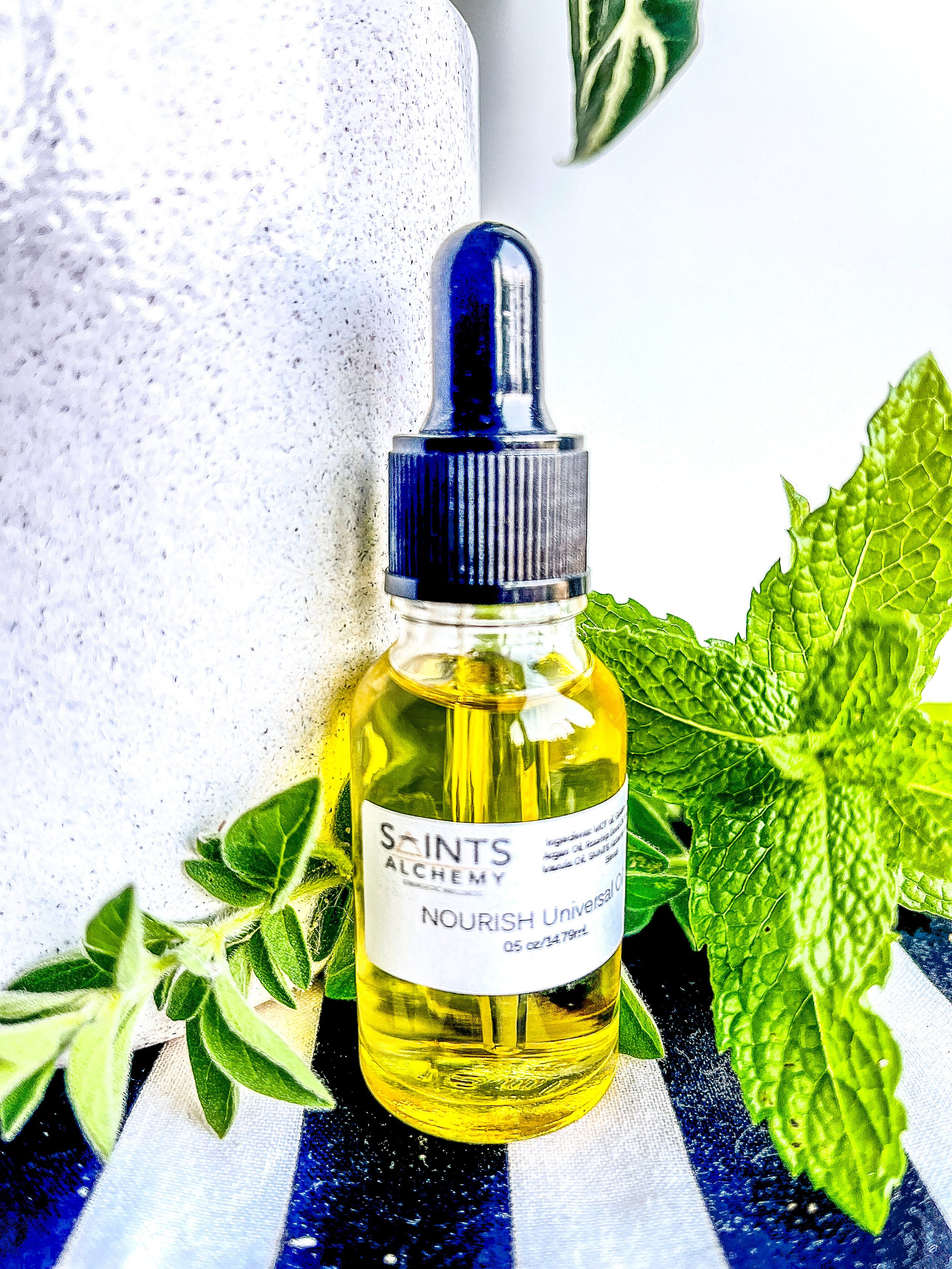 A small glass bottle of yellow essential oil sits among green plants on a cloth with blue, white, and black stripes.