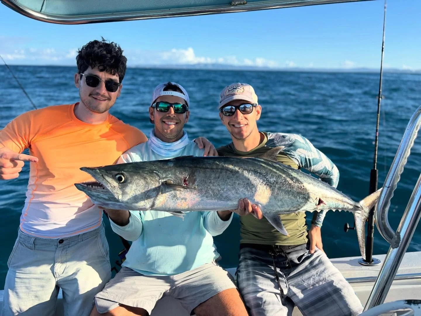 Three anglers on a boat holding a King Mackarel they caught, on top water lures with the ocean and blue sky in the background.