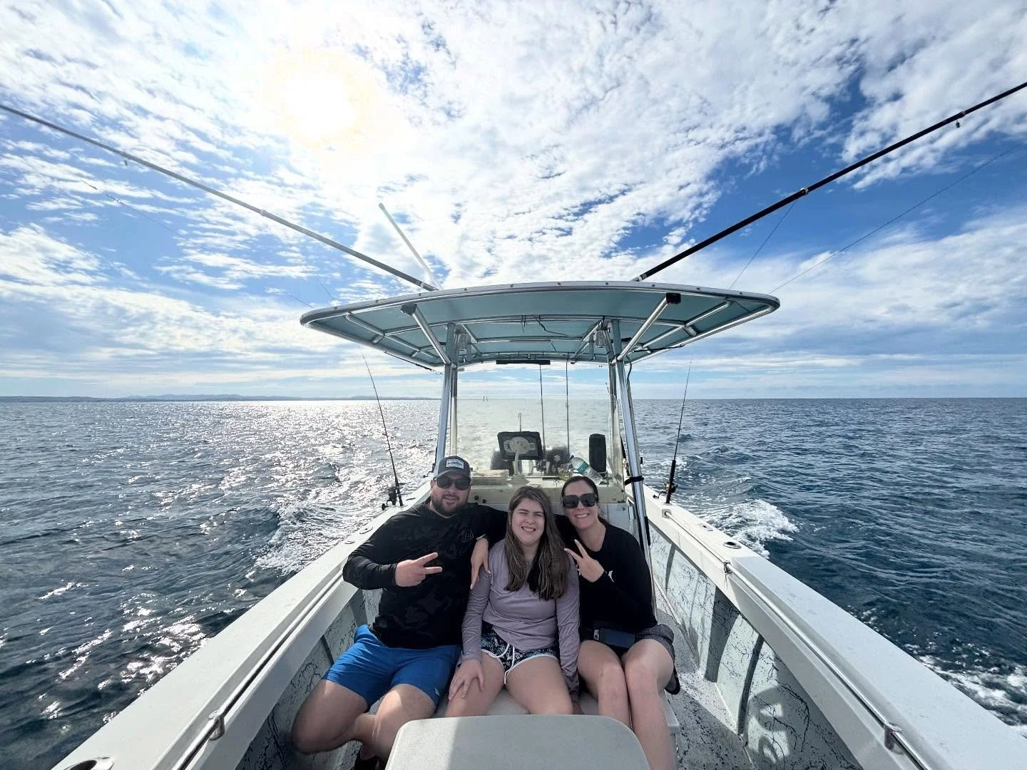 Three people sitting on a boat in the ocean, smiling and posing for the photo with the open sea and partly cloudy sky behind them.