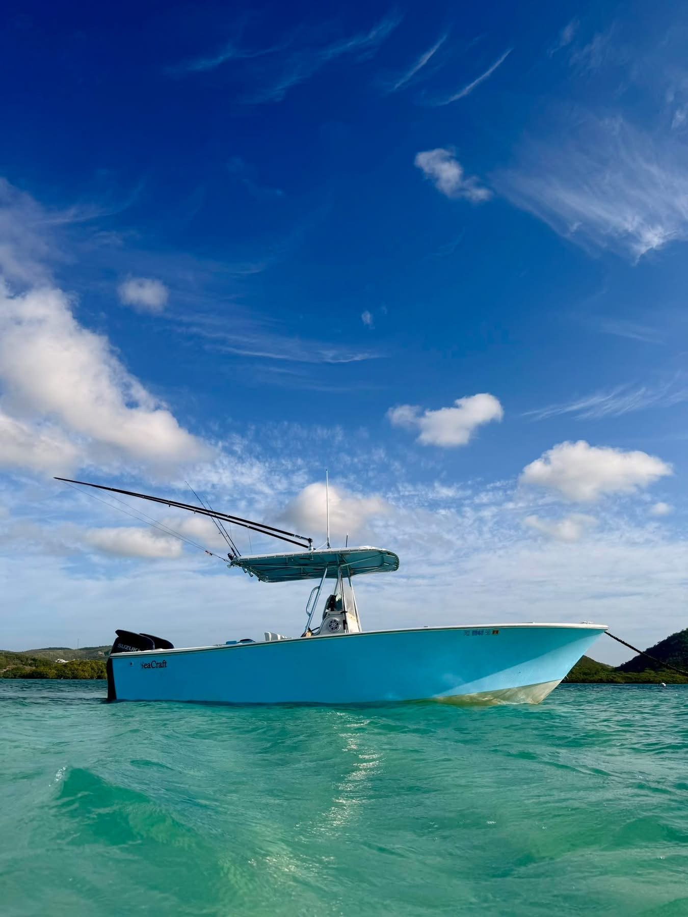 A light blue boat floating on turquoise water with a blue sky and some clouds above and land in the background.