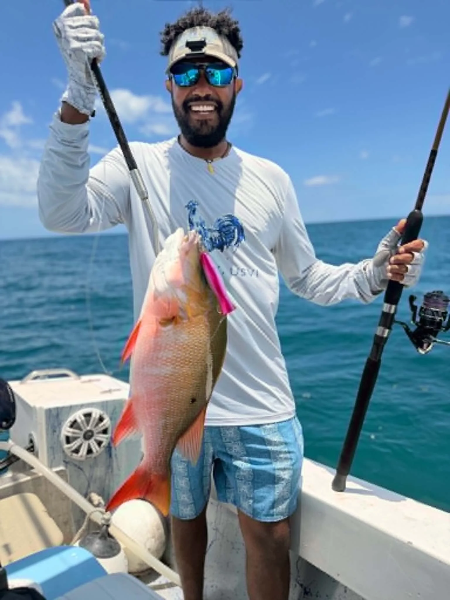 A man standing on a boat holding a fishing rod with a large fish caught on the line. The man is wearing sunglasses, a cap, a long sleeve shirt, and shorts, and is smiling. The scene is set on the ocean under a blue sky with some clouds.
