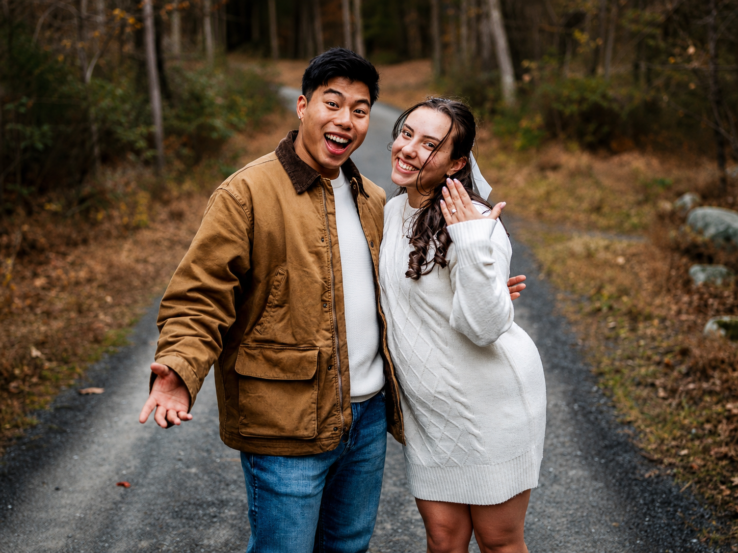 A happy couple standing on a forest trail in autumn, with the woman showing off her engagement ring.