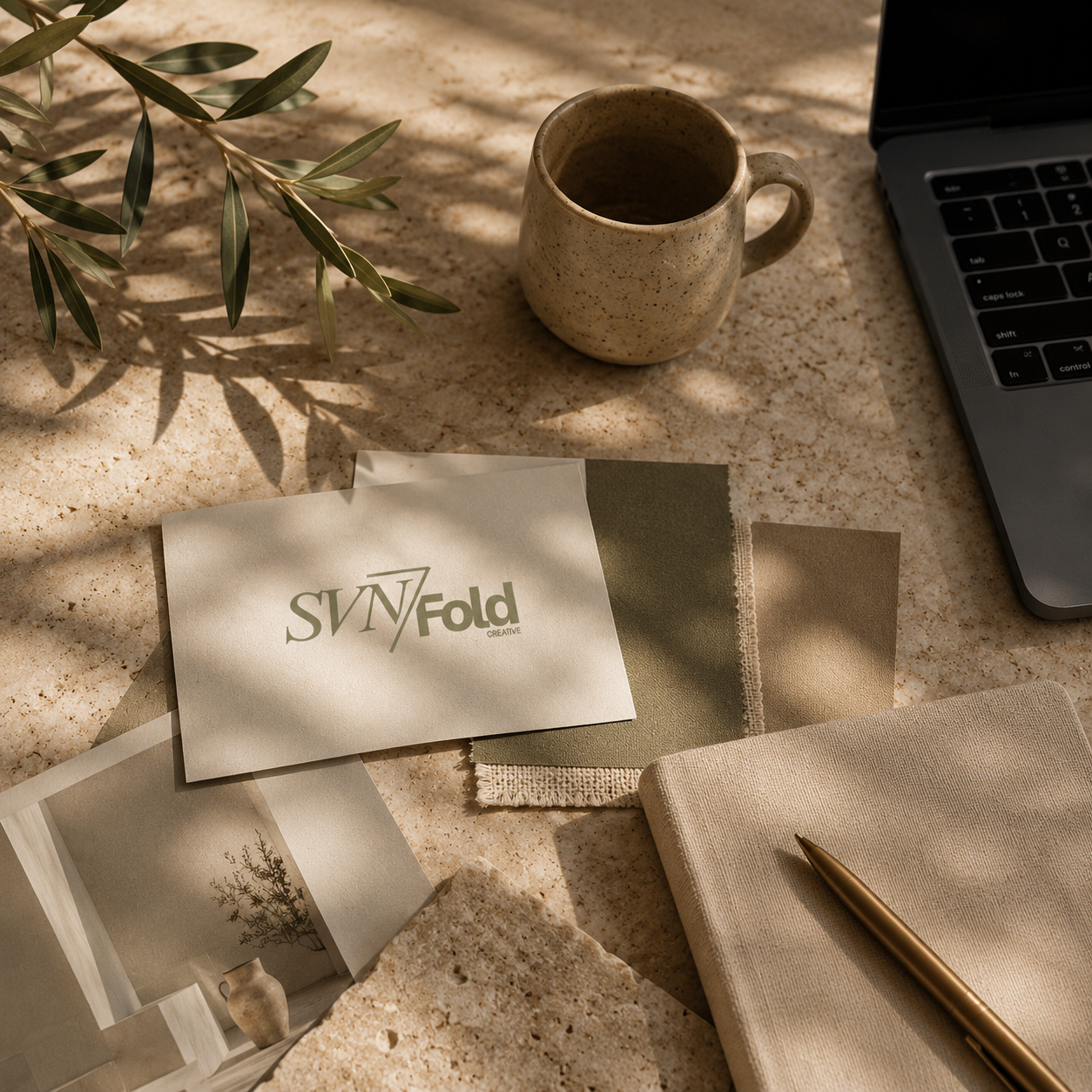 A workspace with a beige notebook, a gold pen, a patterned mug, a laptop, and swatch samples on a speckled cream-colored surface, with shadows and sunlight filtering through a nearby plant.