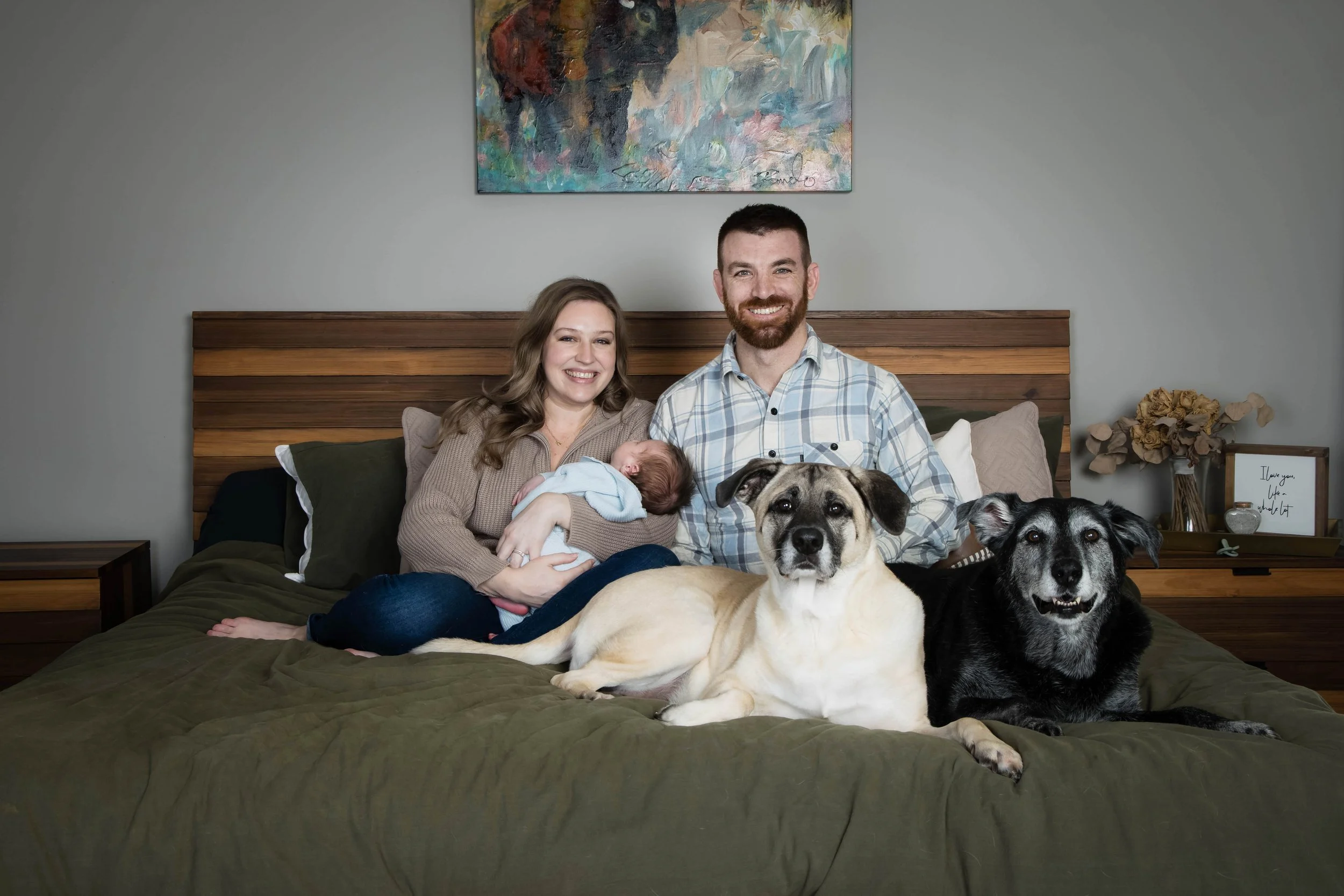 A family of three with two dogs on a bed. The woman is holding a newborn baby. The man has a beard and is wearing a plaid shirt. The family is smiling. The background includes a piece of colorful abstract art, a bedside table with a framed quote, and a vase with dried flowers.