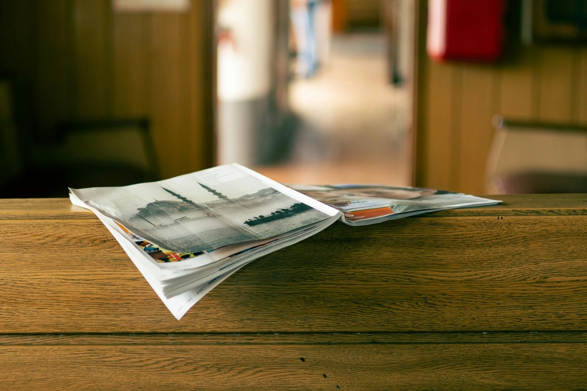 A stack of magazines or newspapers on a wooden table, with the top magazine partially open showing black-and-white photographs of trees and a landscape. The background is blurred with warm indoor lighting.