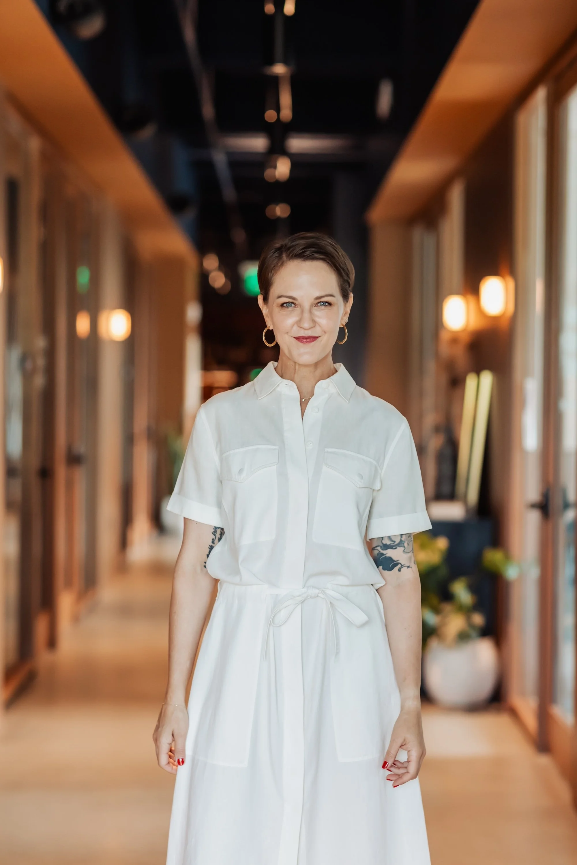 A woman with short brown hair and blue eyes, wearing a white button-up dress with a tied waist, standing in a hallway with warm lighting and wooden accents.