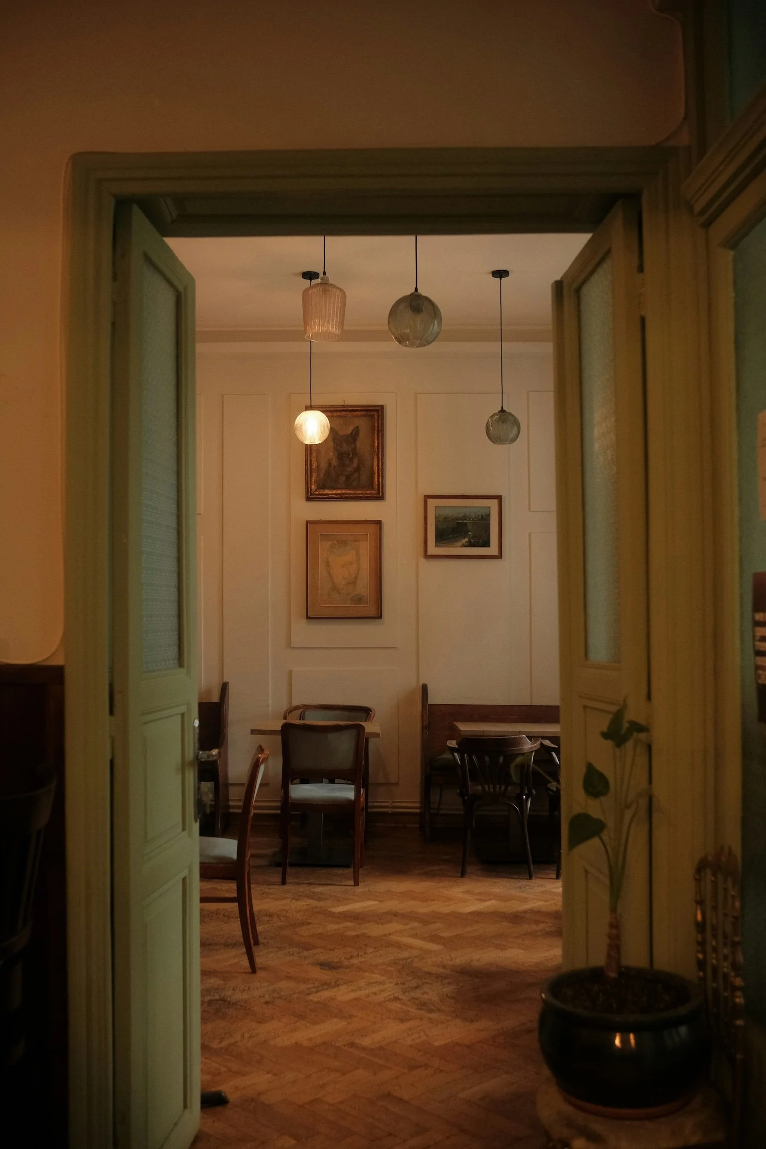 Looking through open green double doors into a cozy, dimly-lit cafe or restaurant interior with wooden tables and chairs, framed artwork on the wall, hanging pendant lights, and a large potted plant on the right.