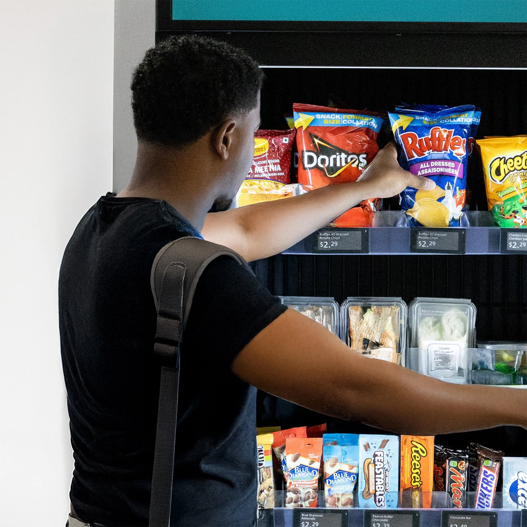 A person with short curly black hair shopping at a snack vending machine, reaching for a bag of Ruffles potato chips.