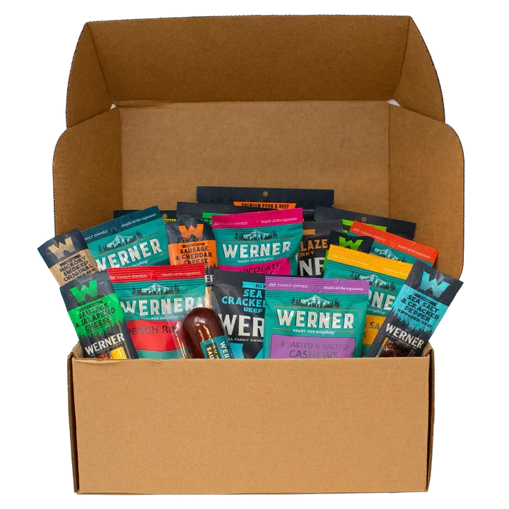 Open cardboard box filled with various Werner snack packages, including flavored cashews, sea crackers, sausage, cheddar cheese snacks, and other mixed nuts.