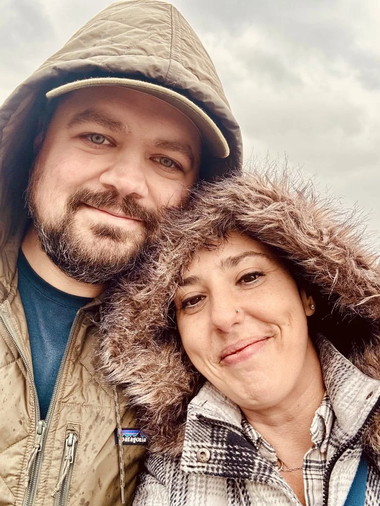 A man and woman outdoors on a cloudy day, both wearing jackets with hoods and smiling at the camera.