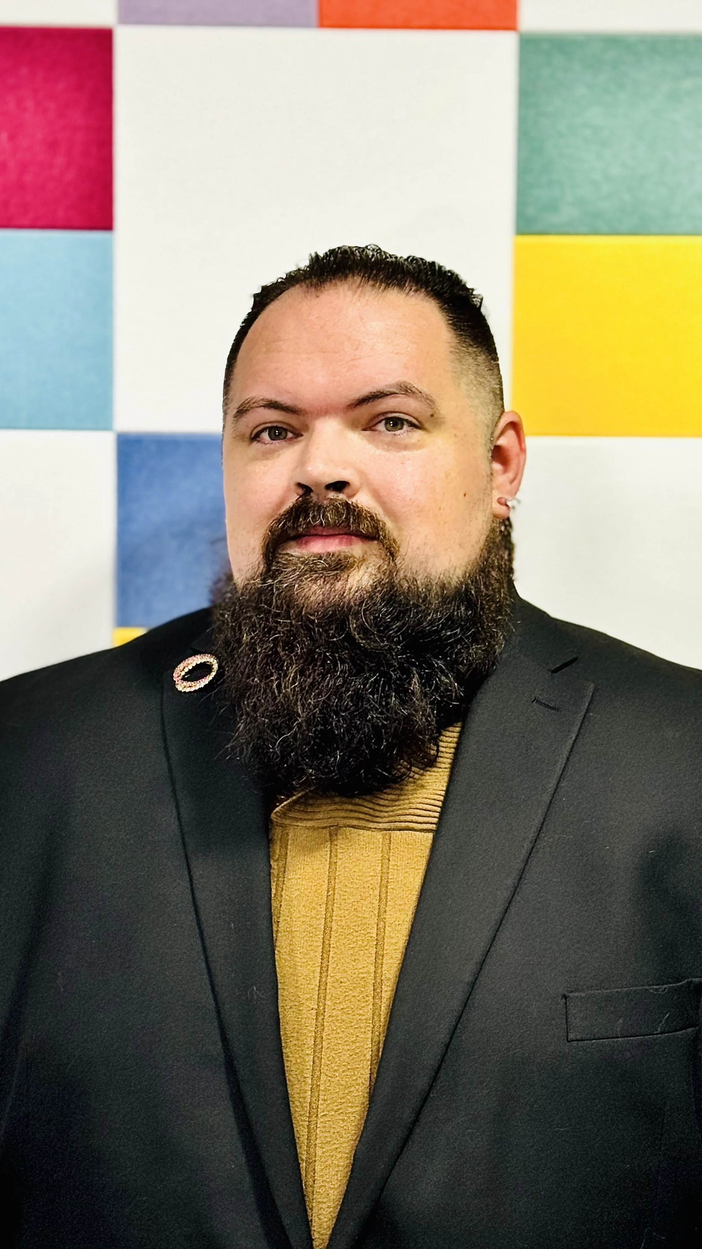 Portrait of a man with a beard and styled hair, wearing a black blazer and a gold-colored sweater, standing in front of a colorful, tiled wall.