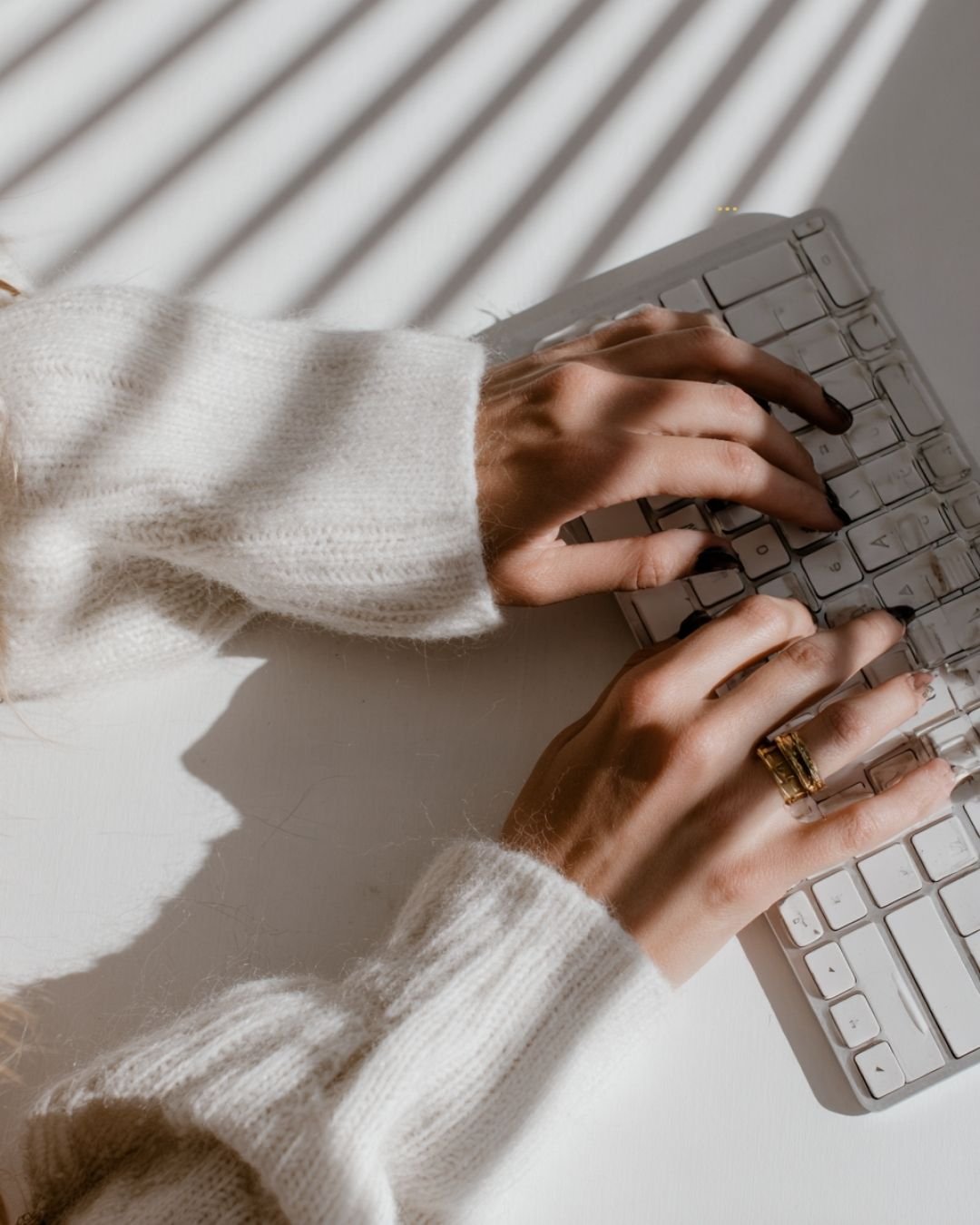 Therapist minh typing on a white keyboard, wearing a white sweater, with sunlight casting shadows.