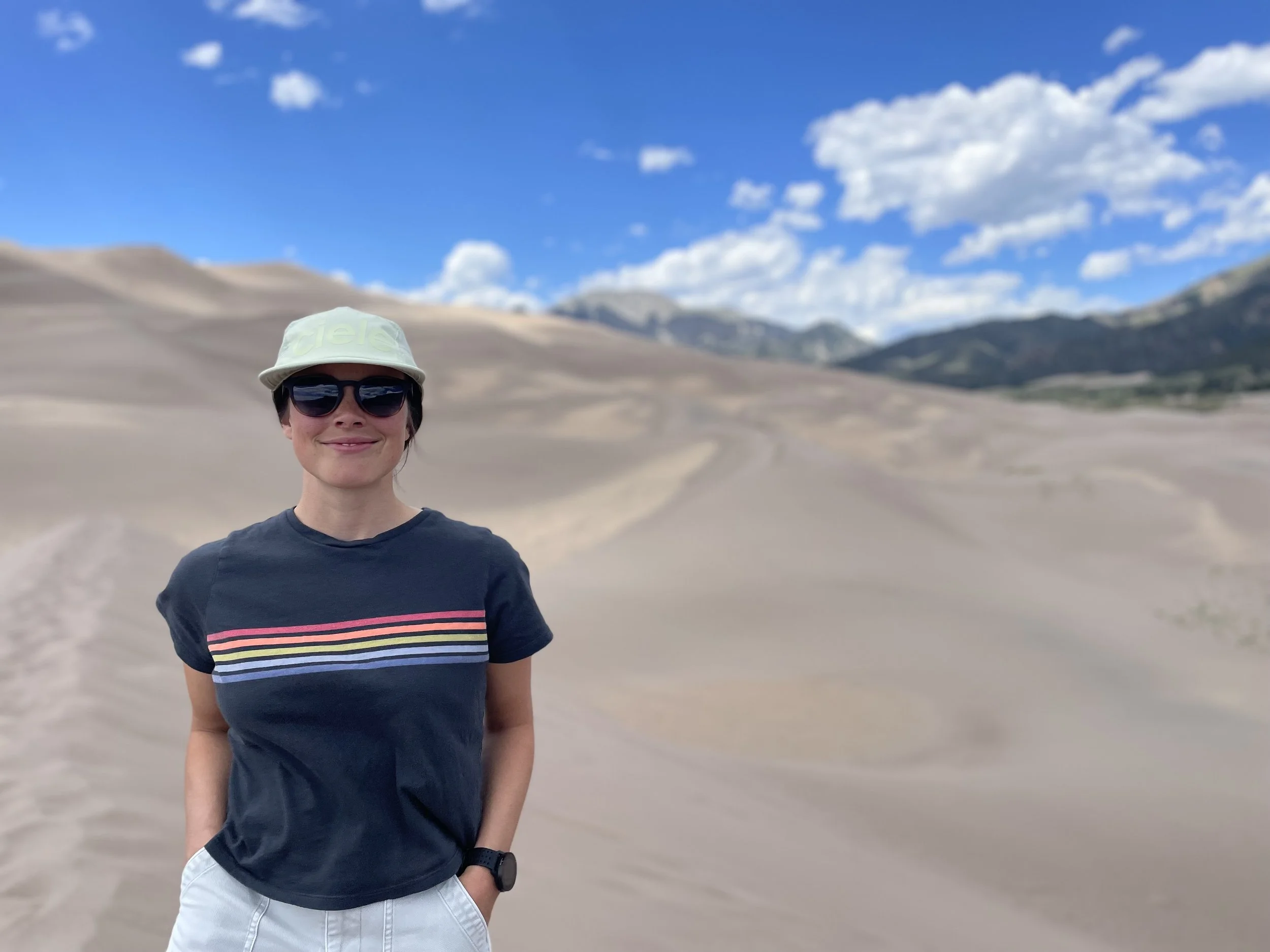 A woman with short, dark hair wearing sunglasses, a light green hat, a navy T-shirt with rainbow stripes, and white shorts, standing outdoors in a desert landscape with sand dunes and mountains in the background under a partly cloudy blue sky.