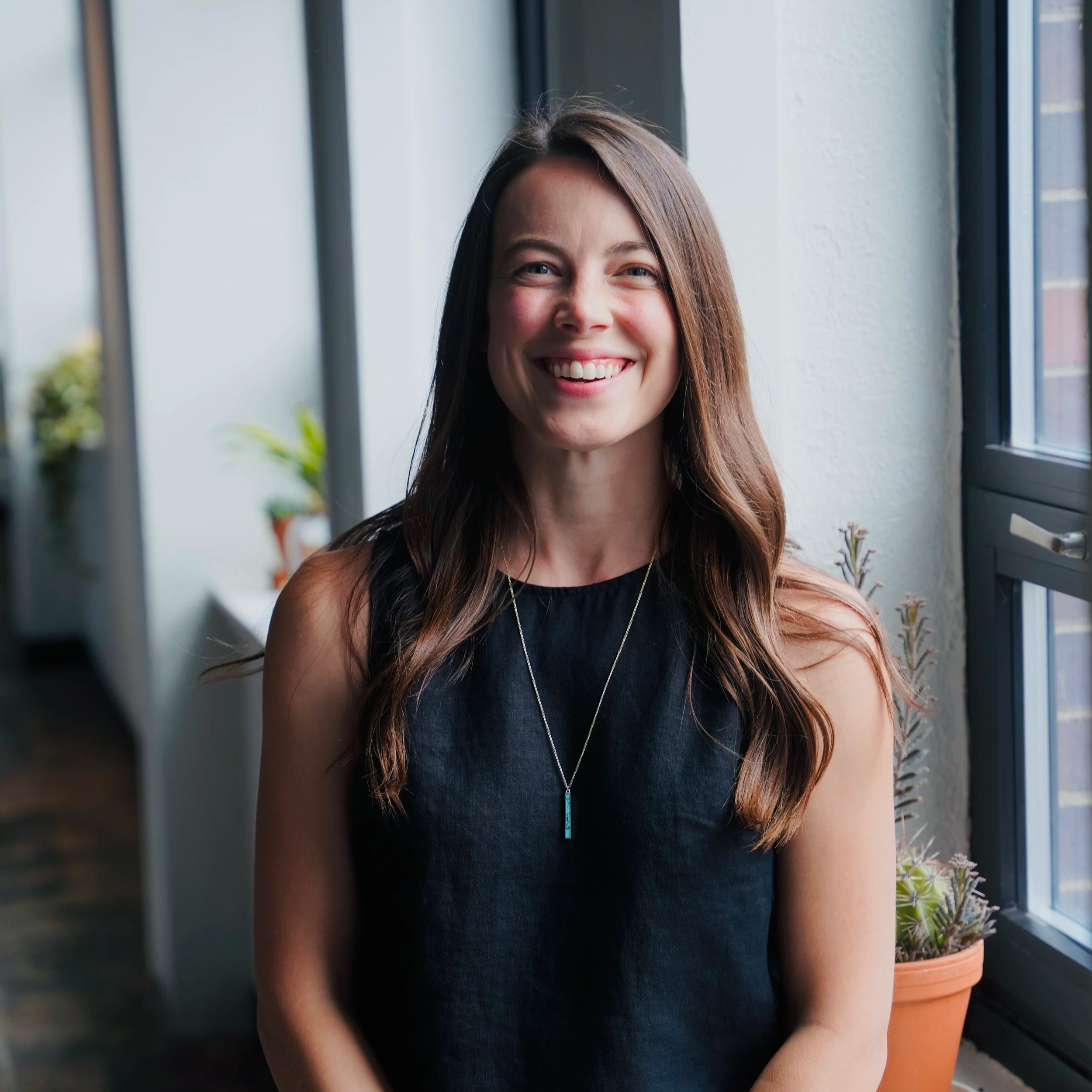 A smiling woman with long brown hair standing by a window indoors, there are plants and flowers in the background.