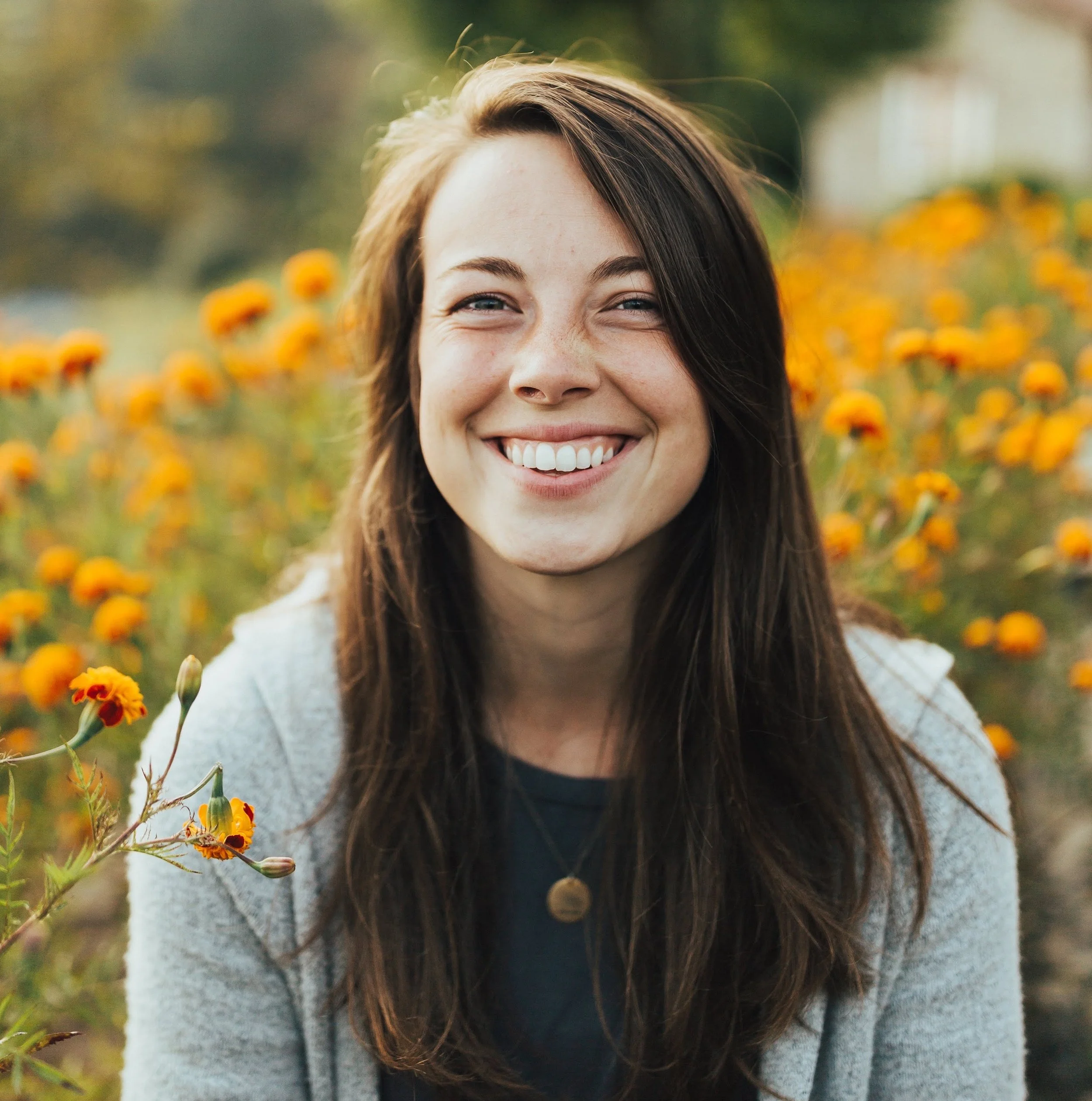 A young woman with brown hair smiling outdoors, surrounded by orange and yellow flowers with a soft-focus background.