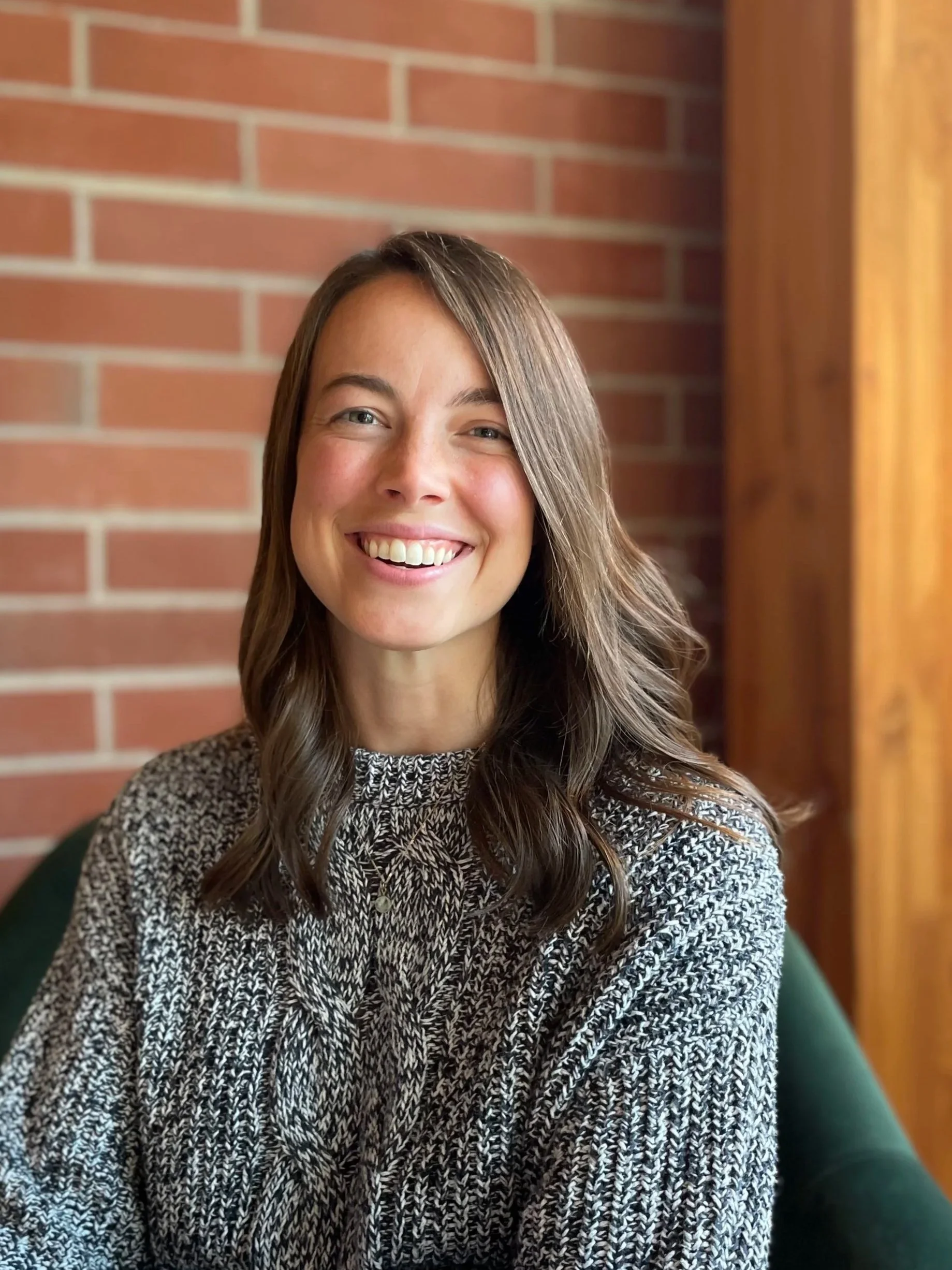 A smiling woman with long wavy brown hair wearing a patterned black and white sweater, sitting on a green chair in front of a brick wall.