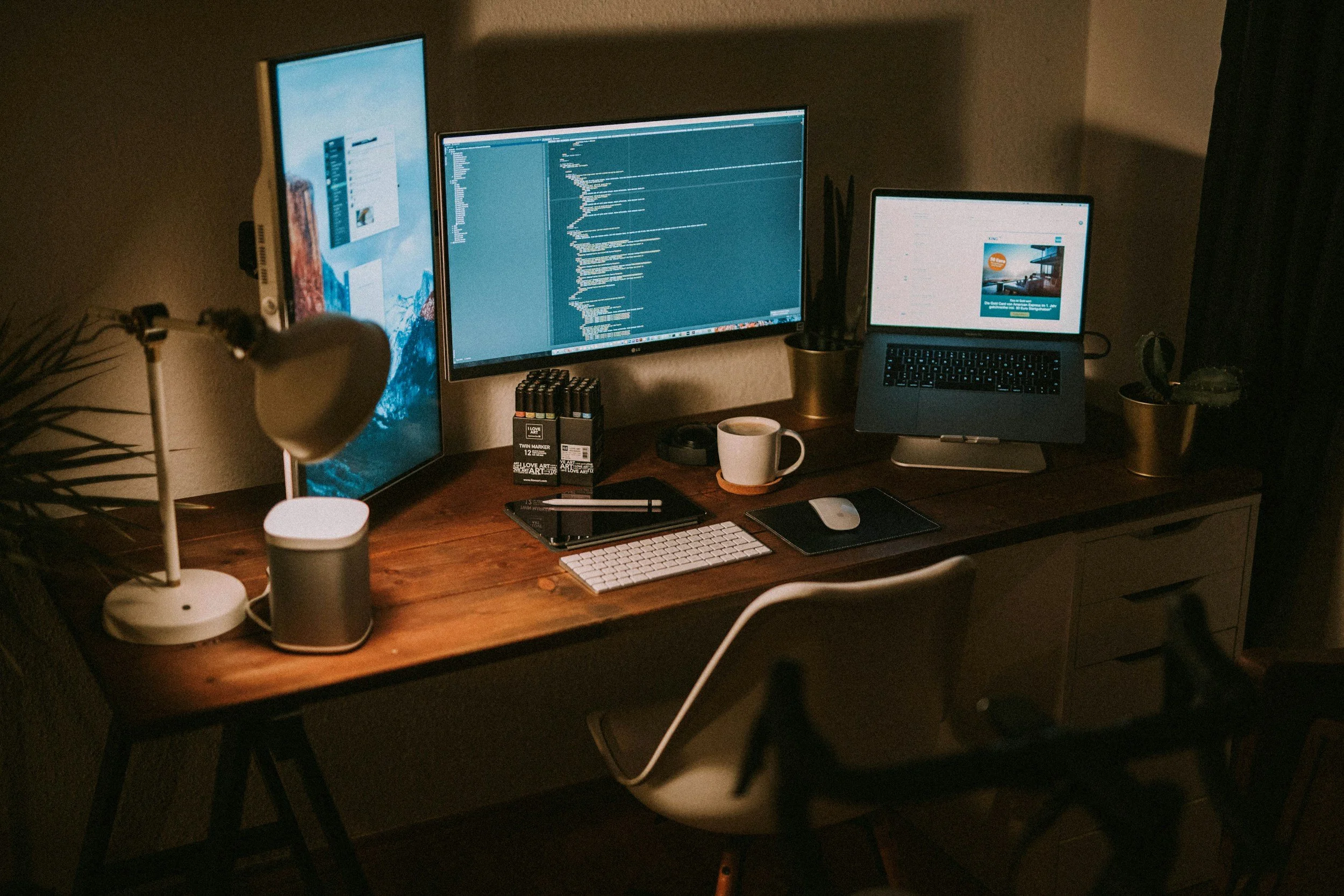 A computer workspace with multiple monitors, a laptop, a keyboard, mouse, coffee mug, and various desk accessories on a wooden desk in a dimly lit room.