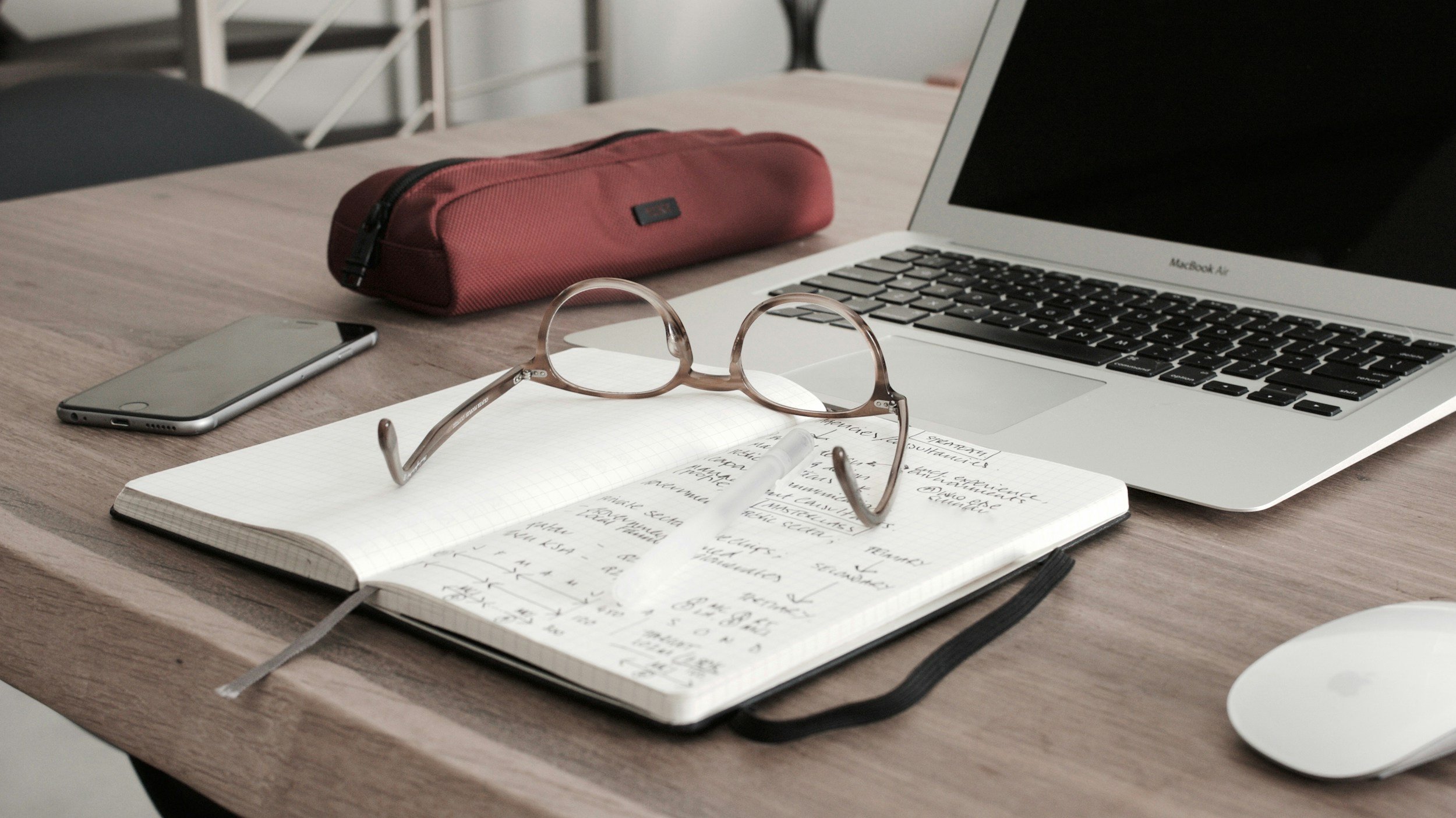 A wooden desk with an open notebook containing handwritten notes, a pair of eyeglasses resting on the notebook, a silver smartphone, a MacBook Air laptop, a maroon pencil case, and a white computer mouse.