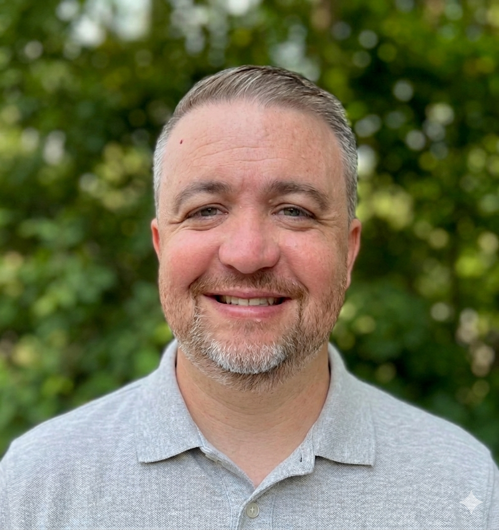 A smiling man with short gray hair, a beard, and blue eyes, wearing a light gray polo shirt, standing outdoors with green foliage in the background.