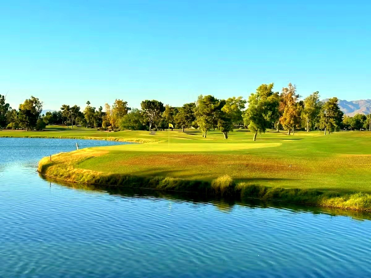 Padre Course at Camelback Golf Club, showing a green beside a lake with a yellow flag, framed by mature trees and distant mountains under a clear blue sky.