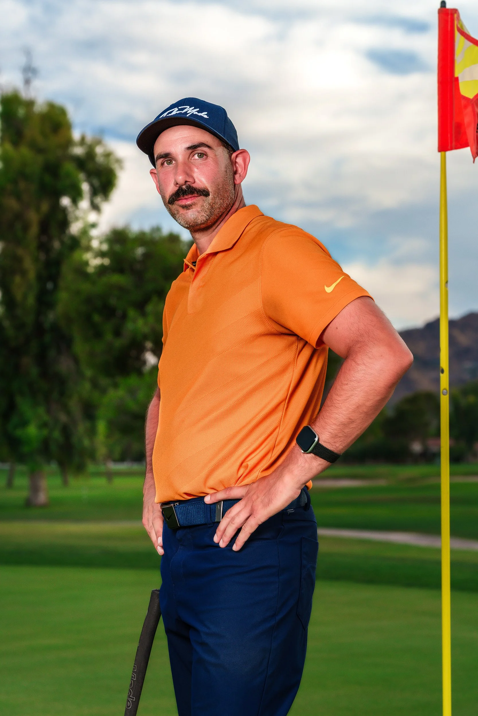 Golf instructor Cody Ceccato standing beside the flagstick on a green at Camelback Golf Club, wearing an orange polo and navy cap.