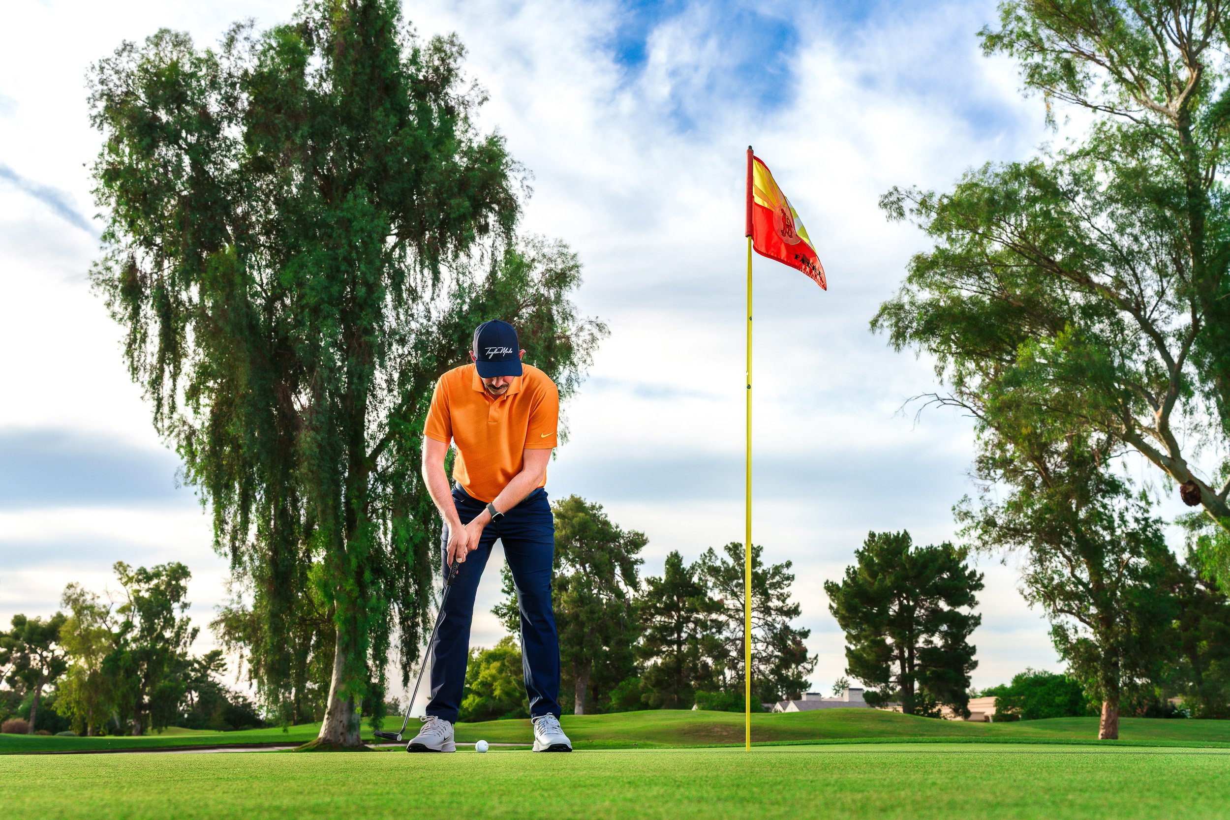 Golfer putting on a green at Camelback Golf Club beside a flagstick, framed by trees and blue sky.