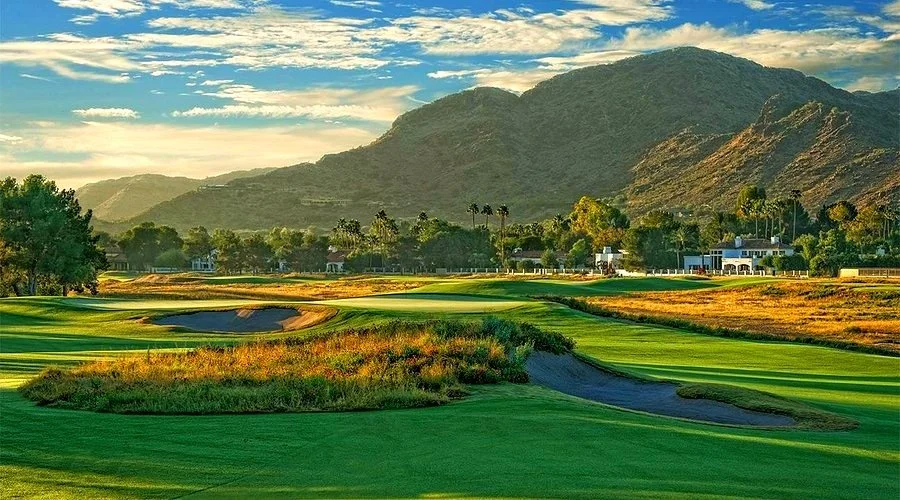 Ambiente Course at Camelback Golf Club, with green fairways and bunkers in the foreground and Camelback Mountain rising behind palm trees and clubhouse buildings at golden hour.