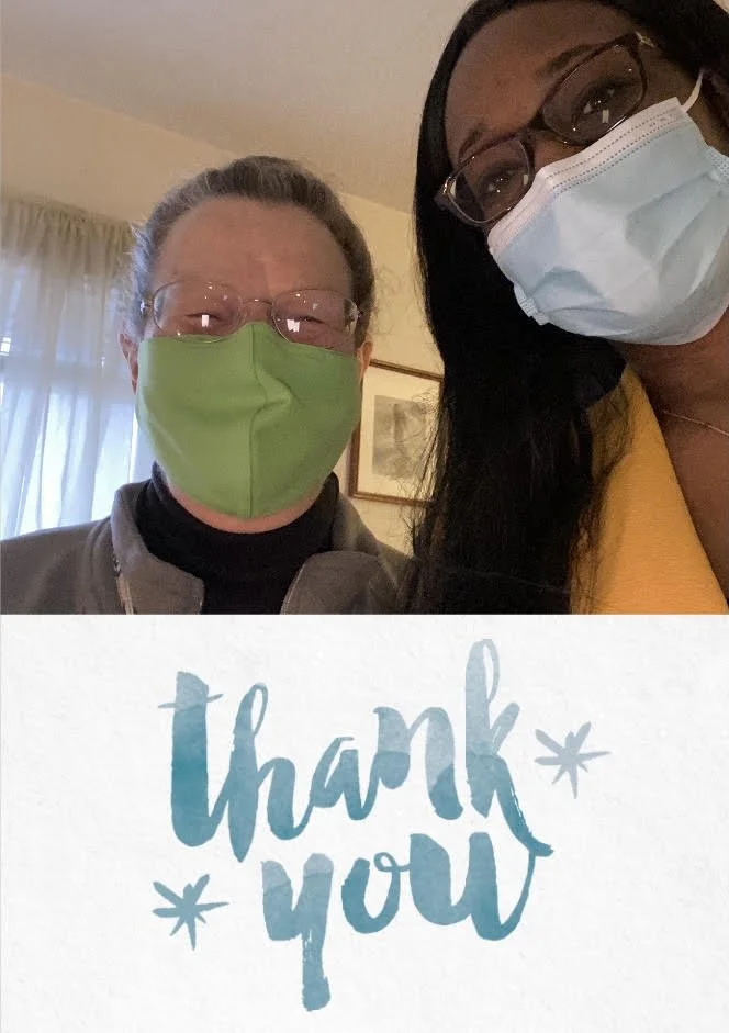 Two women wearing masks, standing indoors, with a handwritten note that says "thank you" seen in front of them.