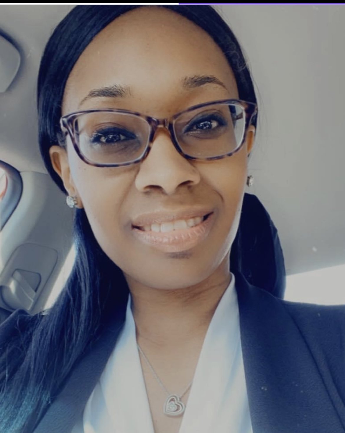 A woman wearing glasses and a black blazer, smiling inside a car.