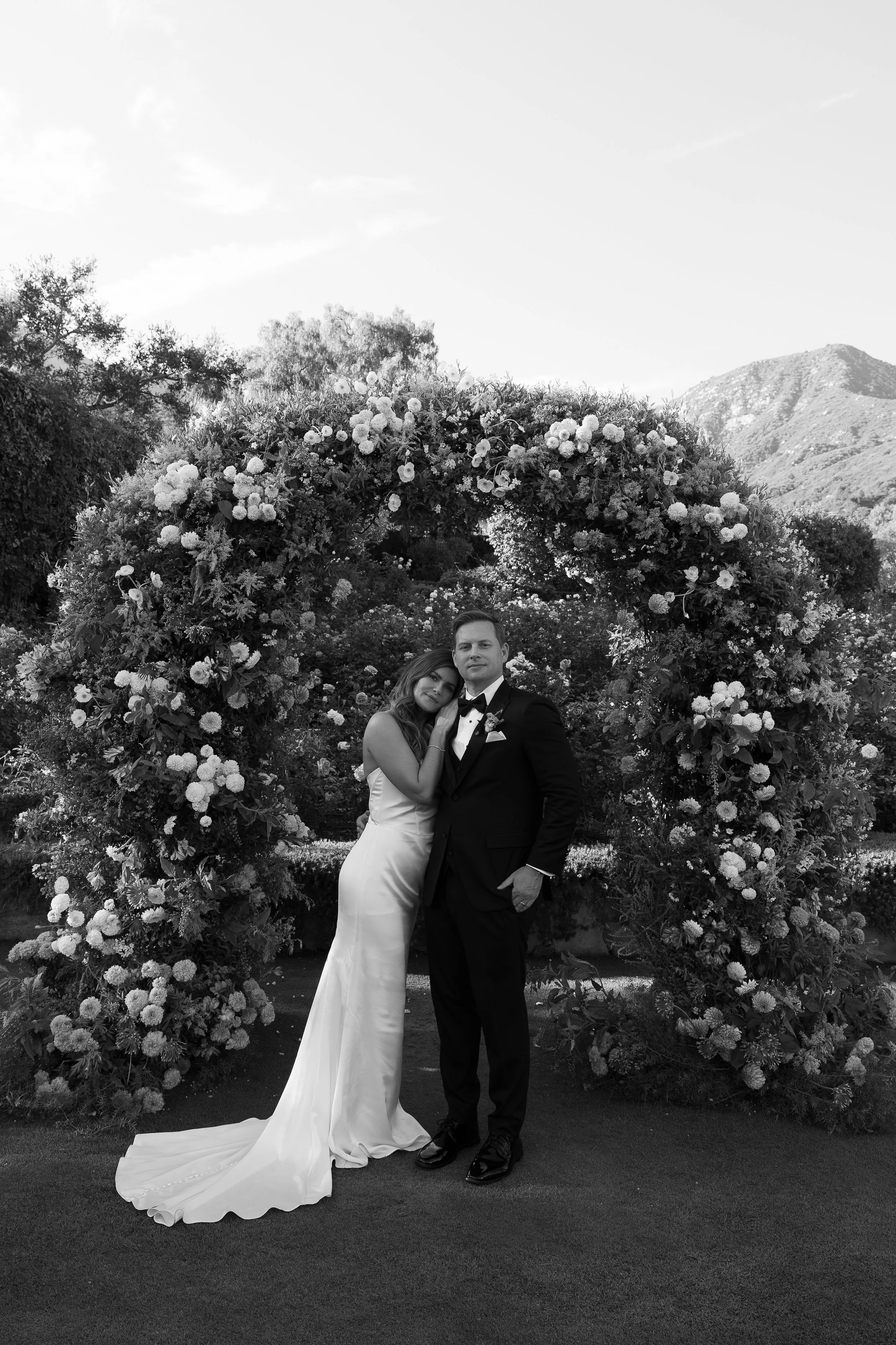 A couple in wedding attire standing in front of a large floral arch with mountains and trees in the background.