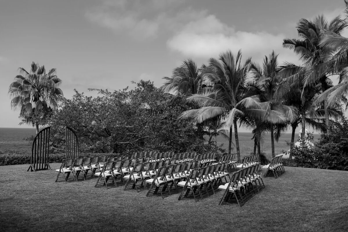 Outdoor wedding ceremony setup with rows of chairs facing a wooden arch, set on grass with palm trees and ocean in the background.