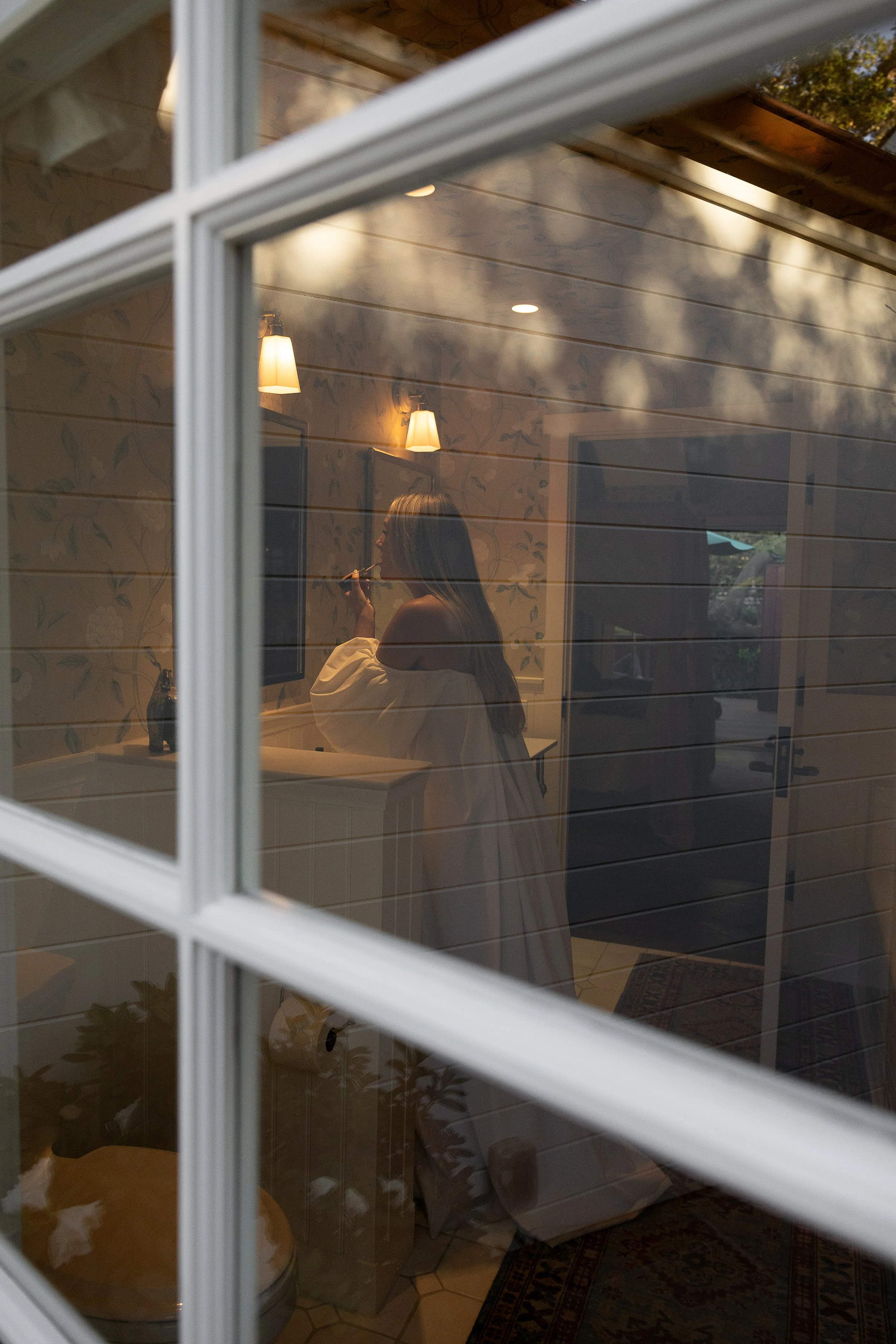 The photo shows a woman with long hair standing inside a room, seen through a window with white grid panes. She is applying makeup or lipstick, wearing an off-the-shoulder top. The room has wooden walls, warm lighting from two wall-mounted lamps, and