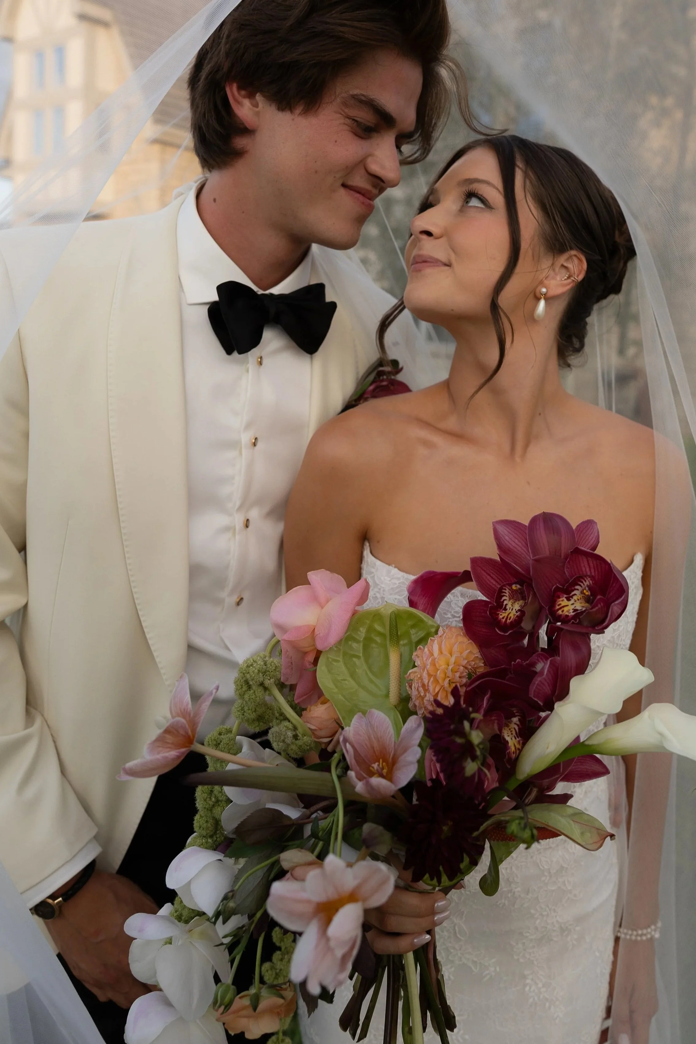 A bride and groom on their wedding day. The groom is dressed in a white tuxedo jacket with a black bow tie and is looking lovingly at the bride. The bride is holding a large bouquet of colorful flowers, including orchids and calla lilies, and is wear