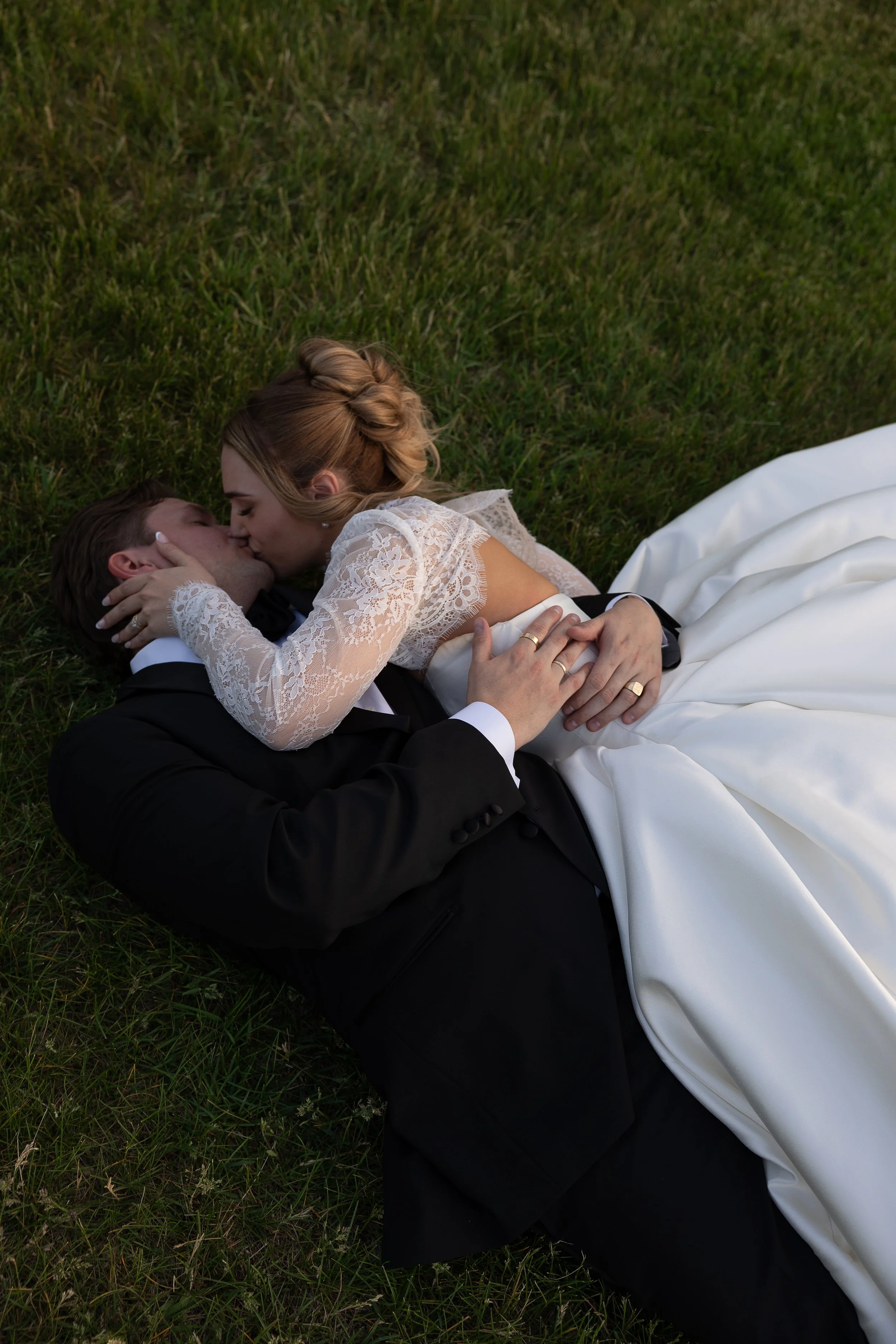 A bride and groom lying on grass, kissing, with the bride in a lace wedding dress and the groom in a black suit.