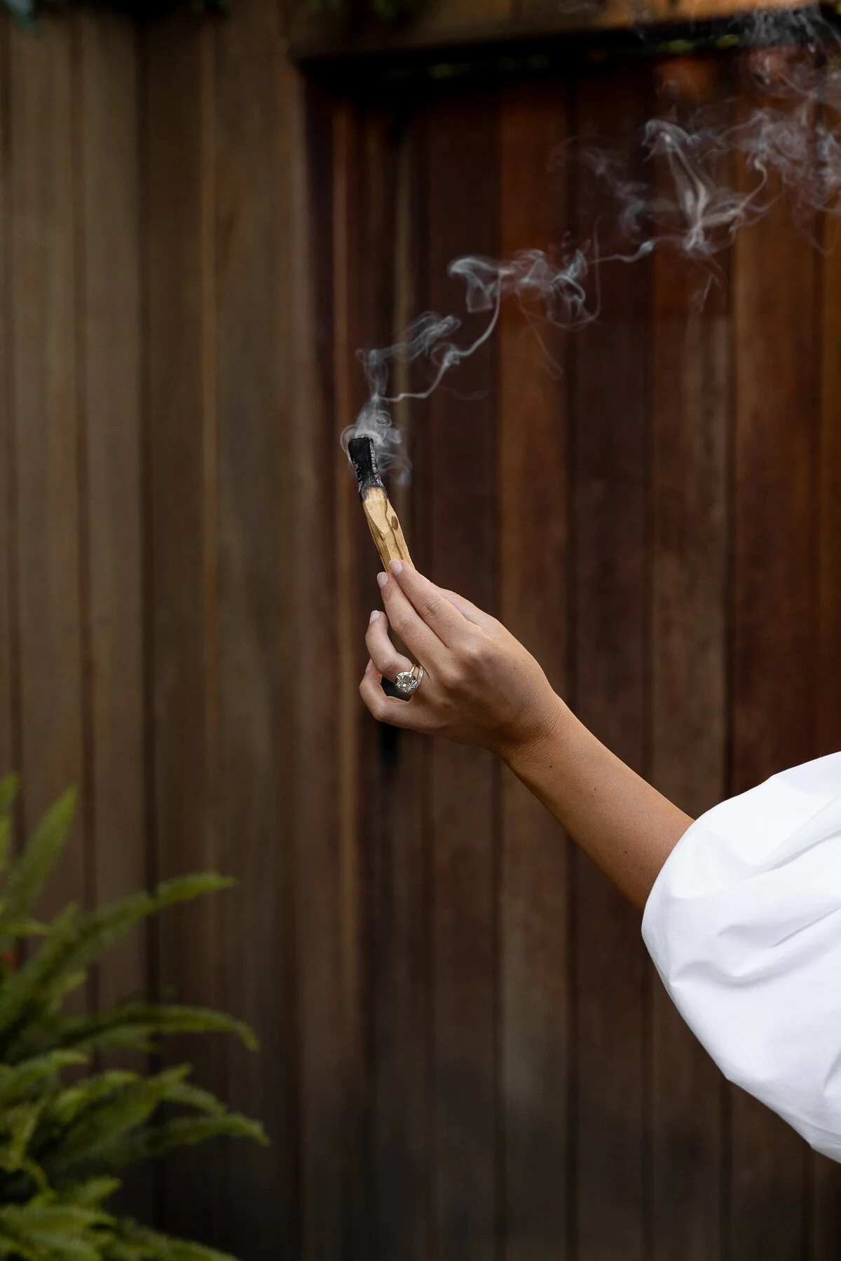 A hand holding a lit matchstick with smoke rising against a wooden fence background.