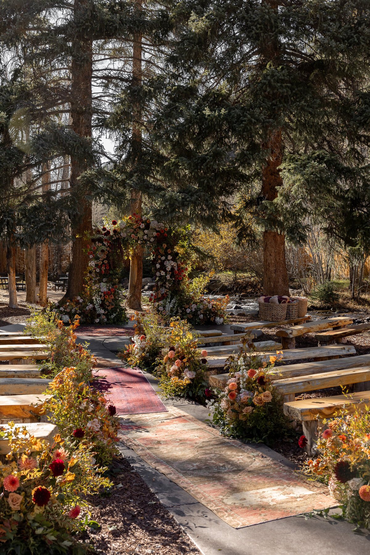 Outdoor wedding setup with floral decorations along a carpeted aisle, arranged between wooden benches and under tall trees with a river in the background.