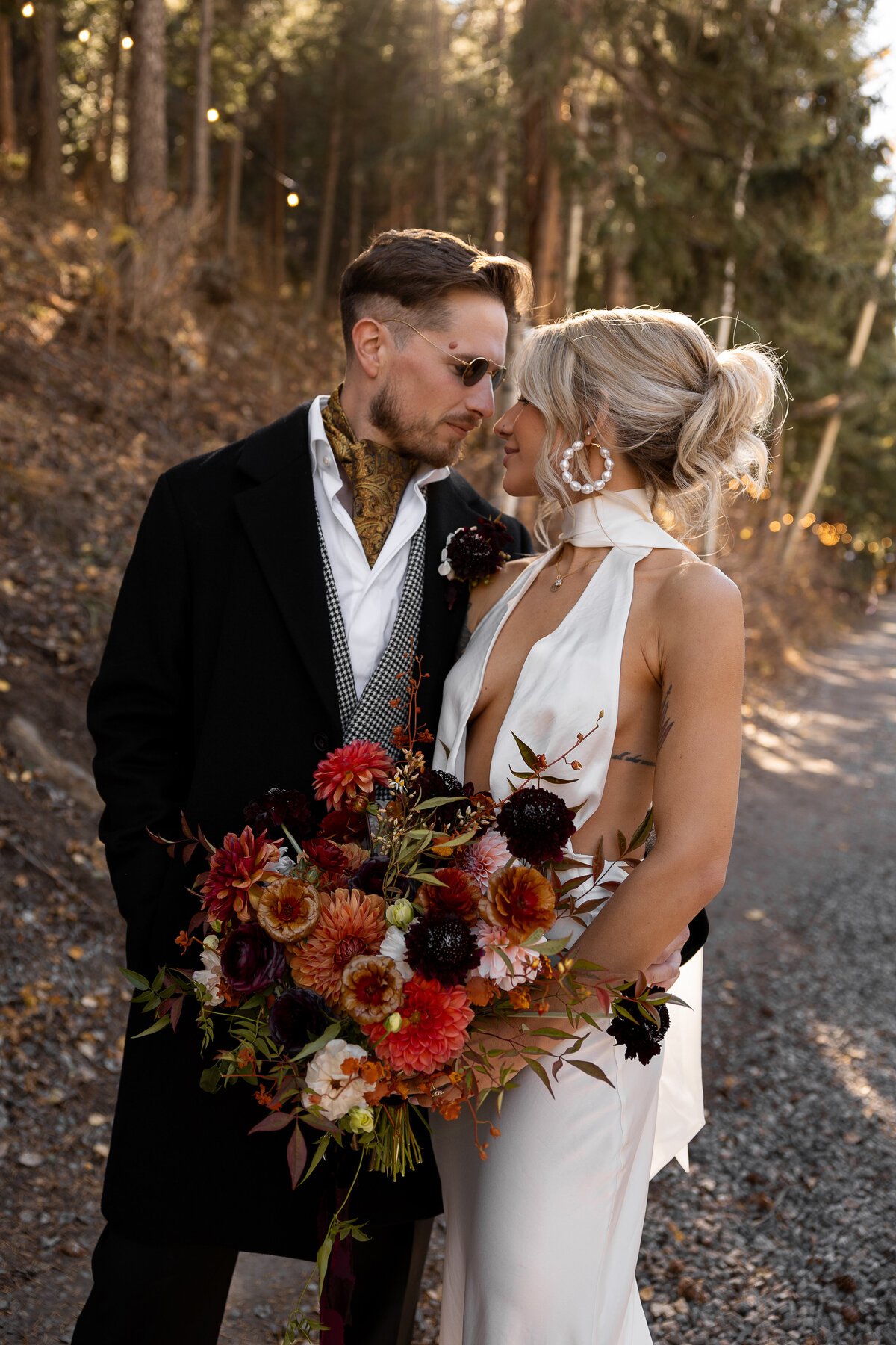 A couple dressed in wedding attire, standing close together outdoors in a wooded area, with sunlight filtering through trees. The woman is holding a large bouquet of dark and bright flowers and wearing a sleeveless, deep neckline white wedding dress.