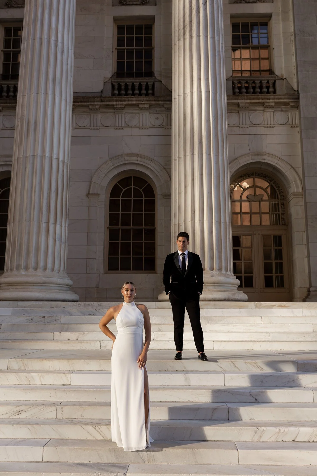 A man and woman dressed in formal attire standing on the steps of a large classical building with marble columns and large windows. The woman is wearing a white dress with a high slit, and the man is wearing a black suit and tie.