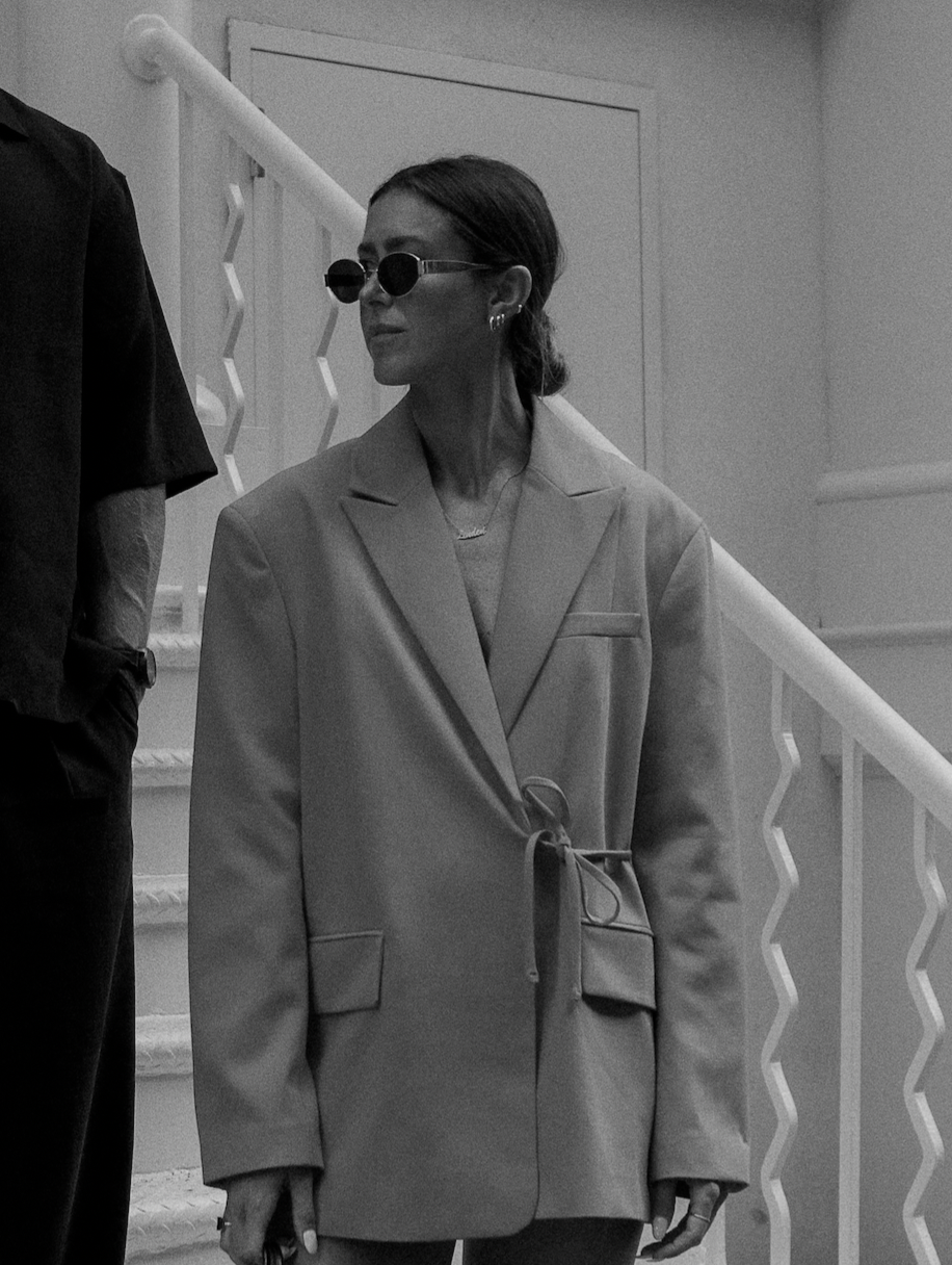 A woman with dark hair tied back, wearing sunglasses, a blazer with a waist tie, and earrings, standing on a staircase interior.