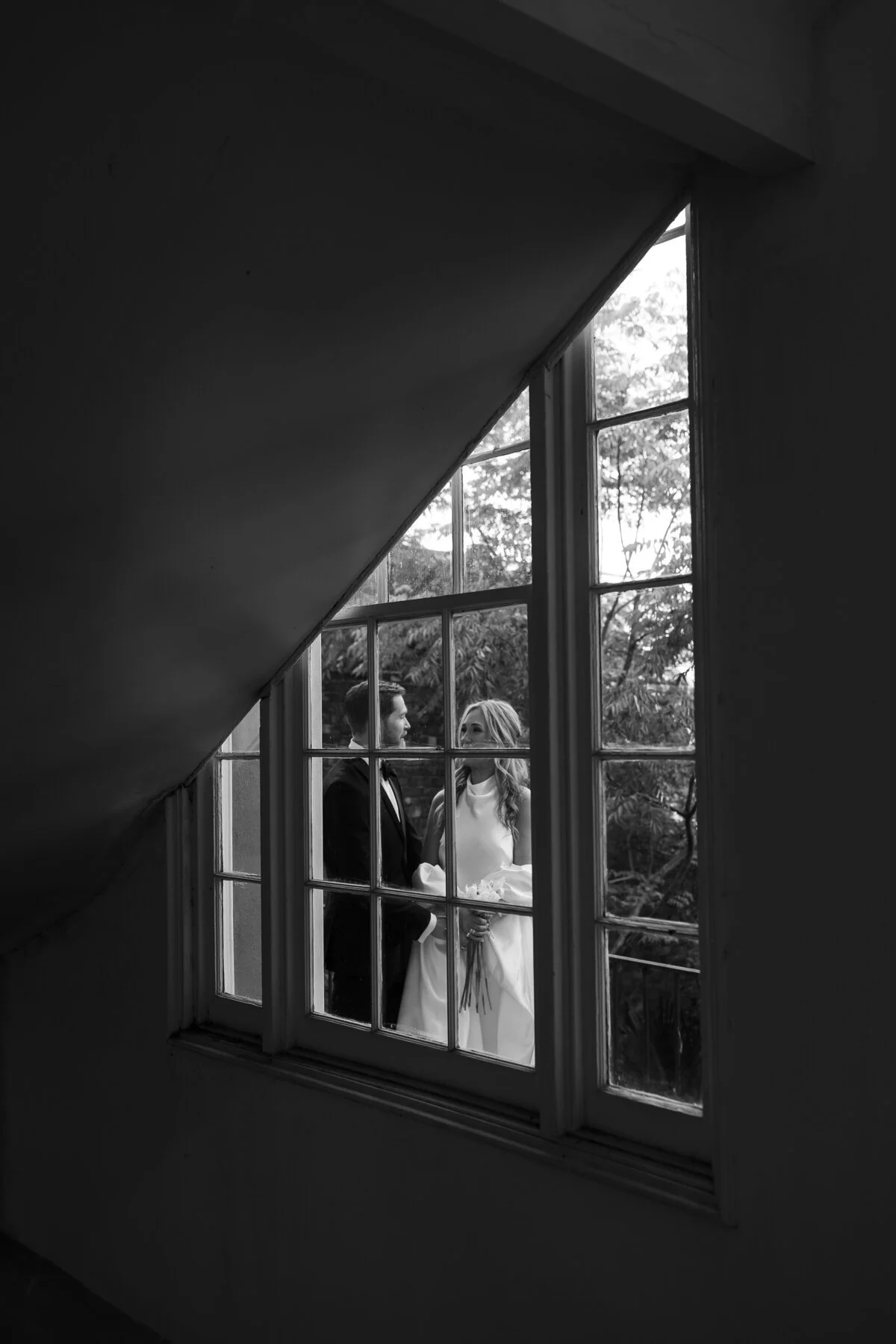A black and white photo of a bride and groom standing outdoors, viewed through a window with multiple panes. The couple is looking at each other, with the bride holding a bouquet.