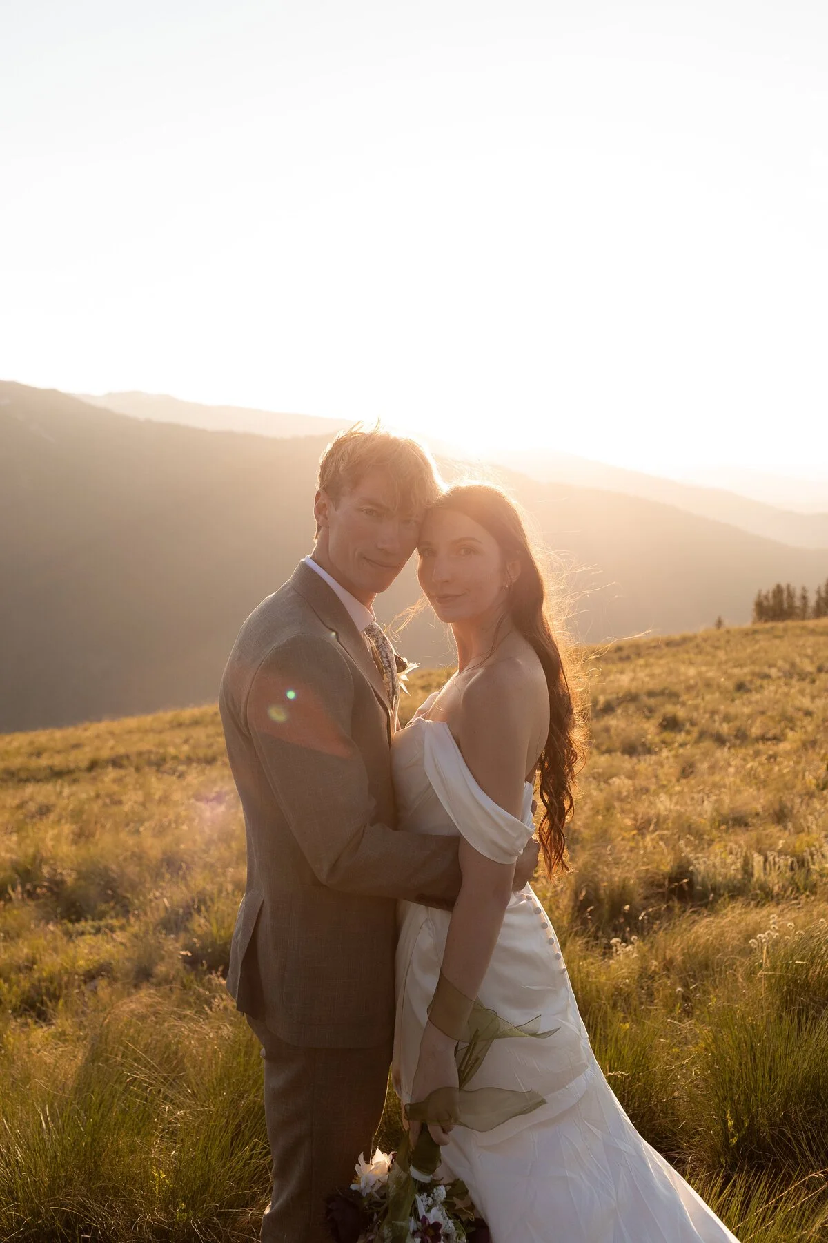 A bride and groom standing close together outdoors during sunset, with mountains in the background. The groom is wearing a gray suit and the bride is in a white wedding dress, holding a bouquet of flowers.