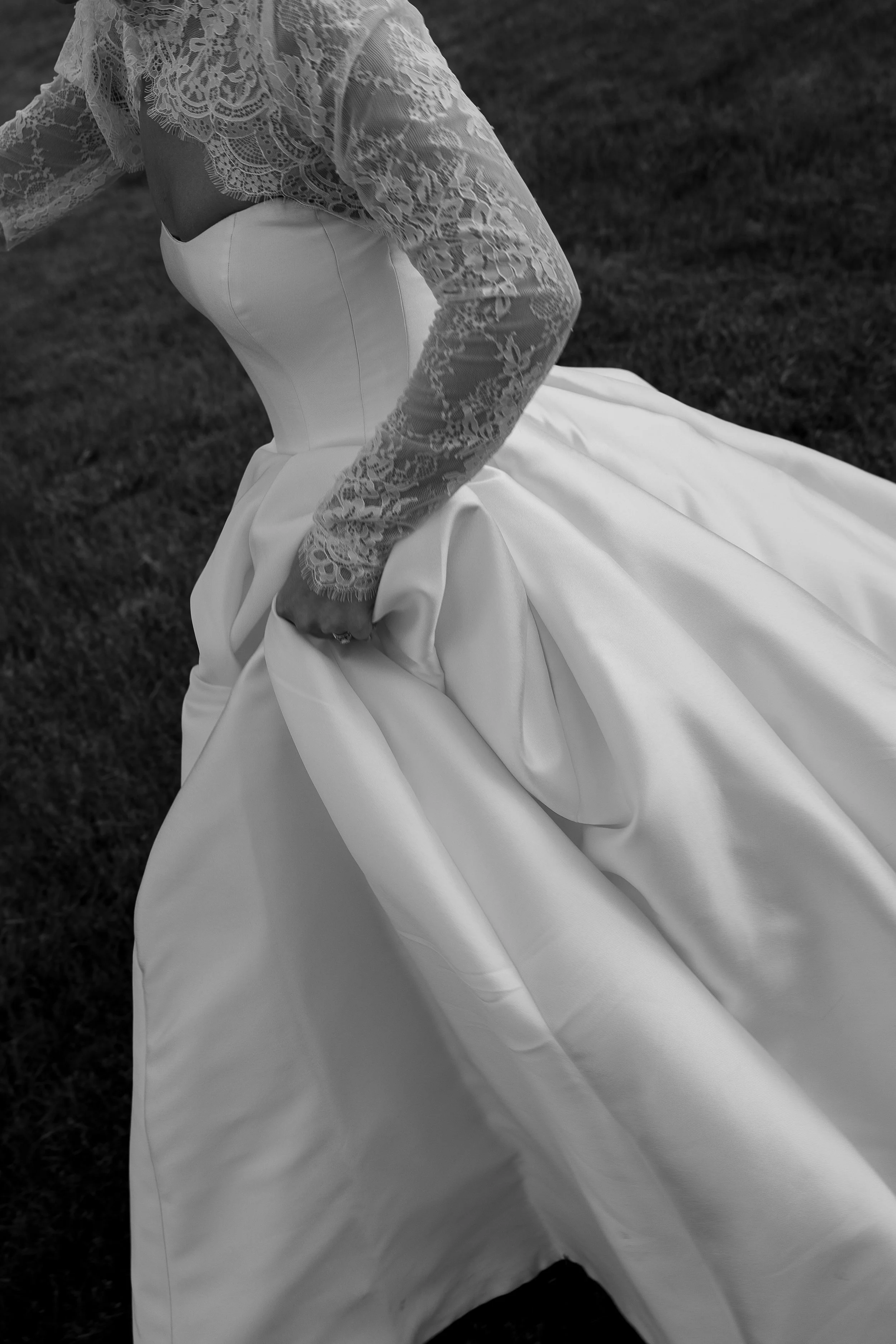 Close-up of a bride wearing a lacy long-sleeve top and a satin wedding dress, bending over outdoors on grass.
