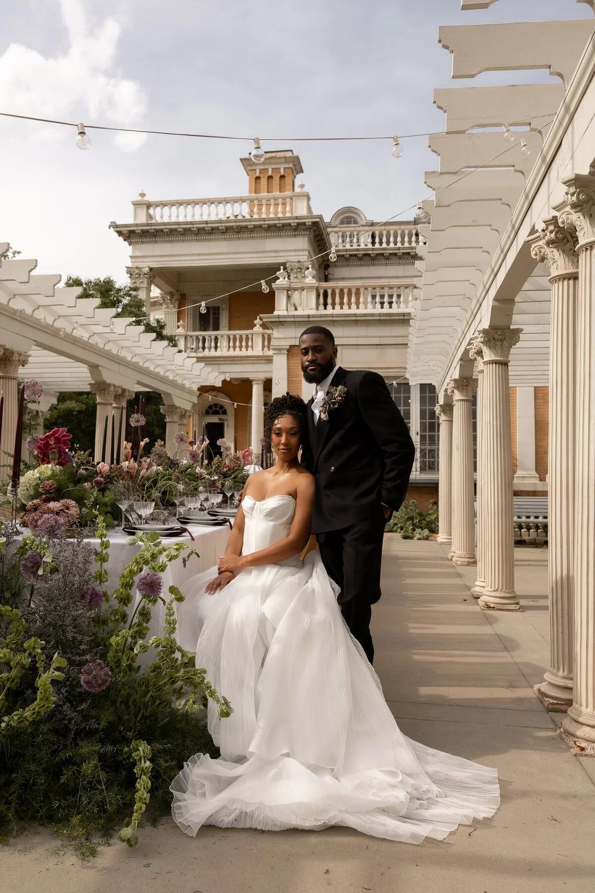 A bride in a white strapless wedding dress sits on a chair, and a groom in a black tuxedo stands beside her, in an outdoor estate setting with classical columns, a decorated table, and floral arrangements.