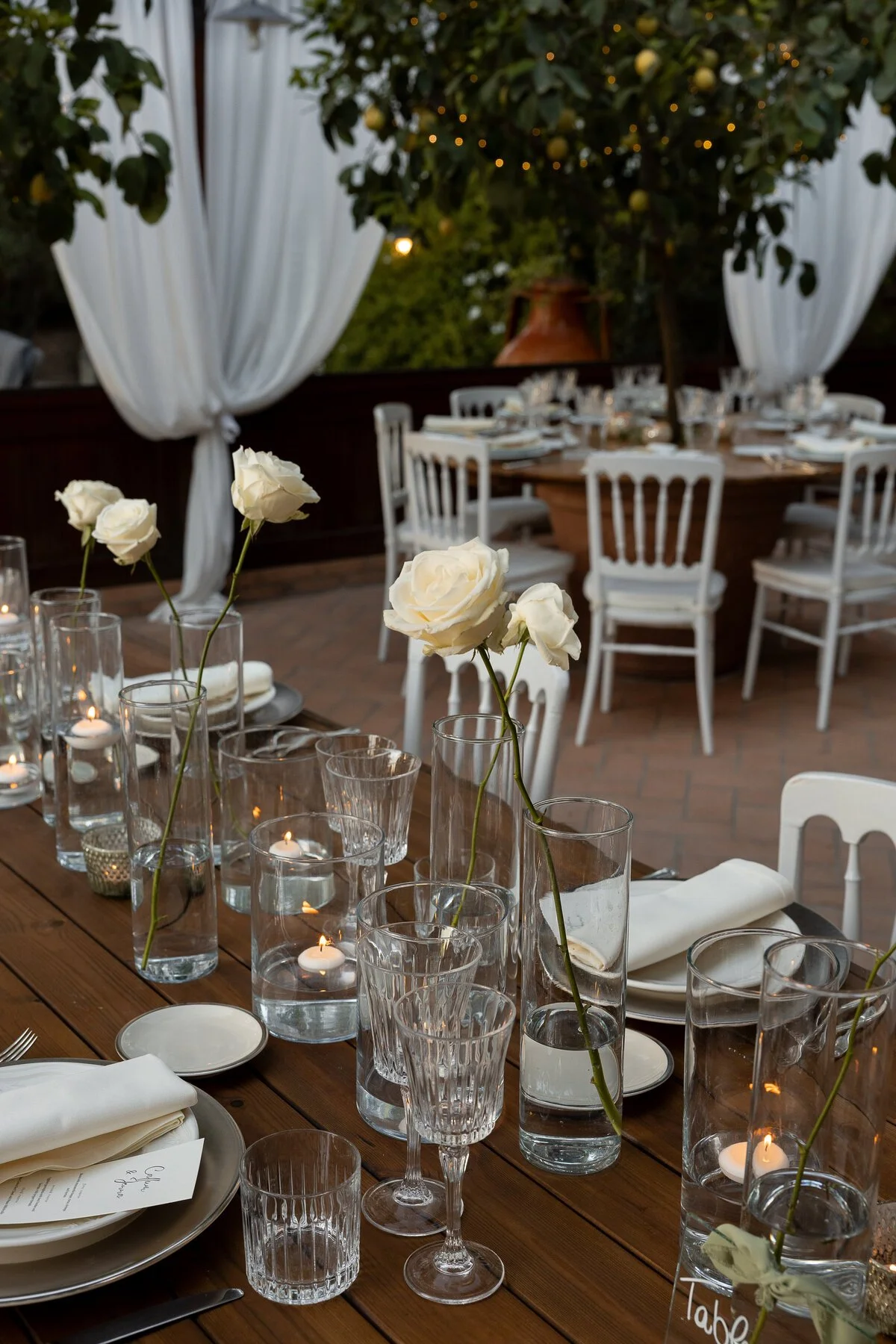 Table decorated for a formal event with white roses in tall glass vases, lit candles in small glass holders, white plates, napkins, and glassware. In the background, a circular table with white chairs and a large potted lemon tree with yellow lemons.