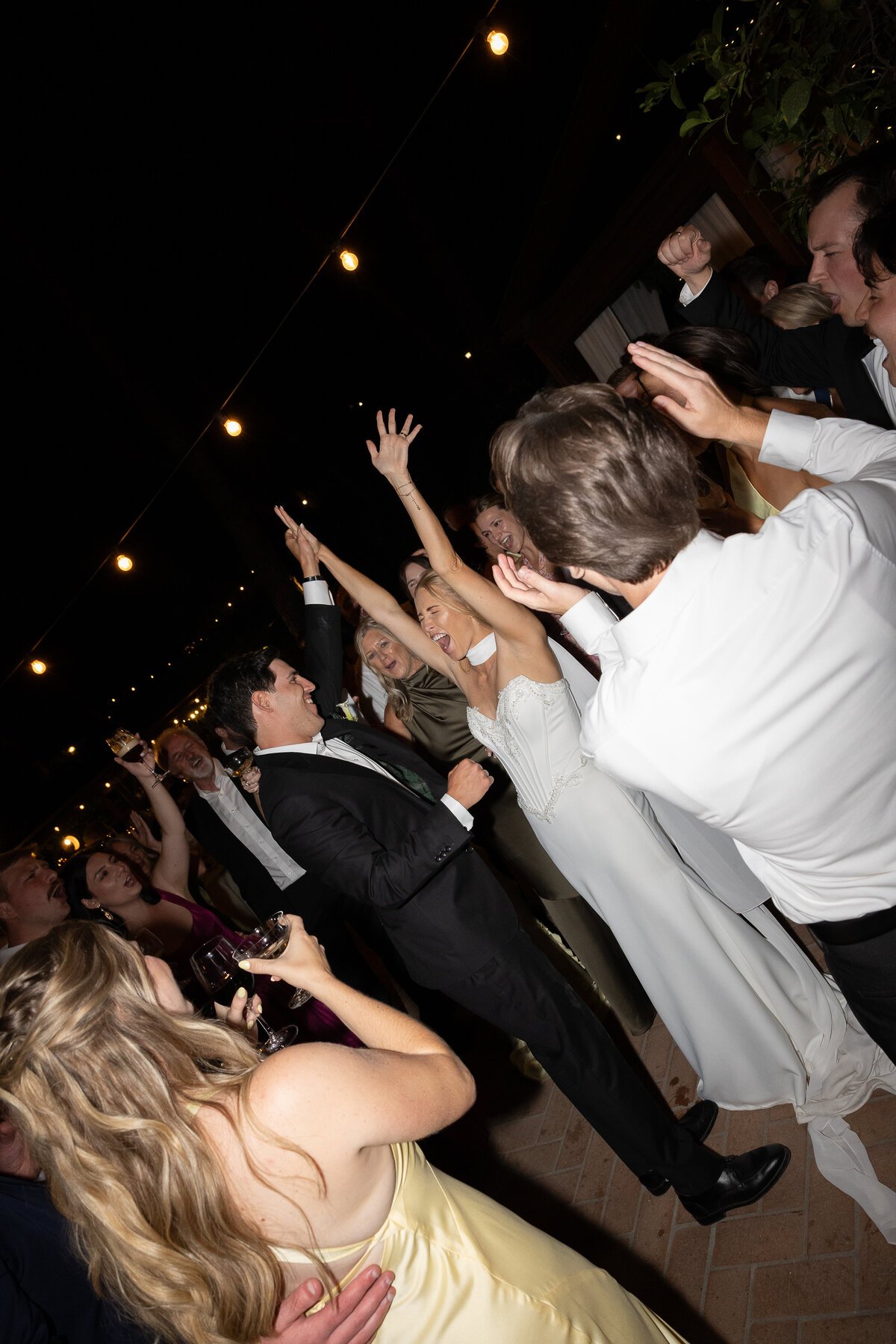 Group of people celebrating at a wedding reception, dancing and smiling under string lights at night.