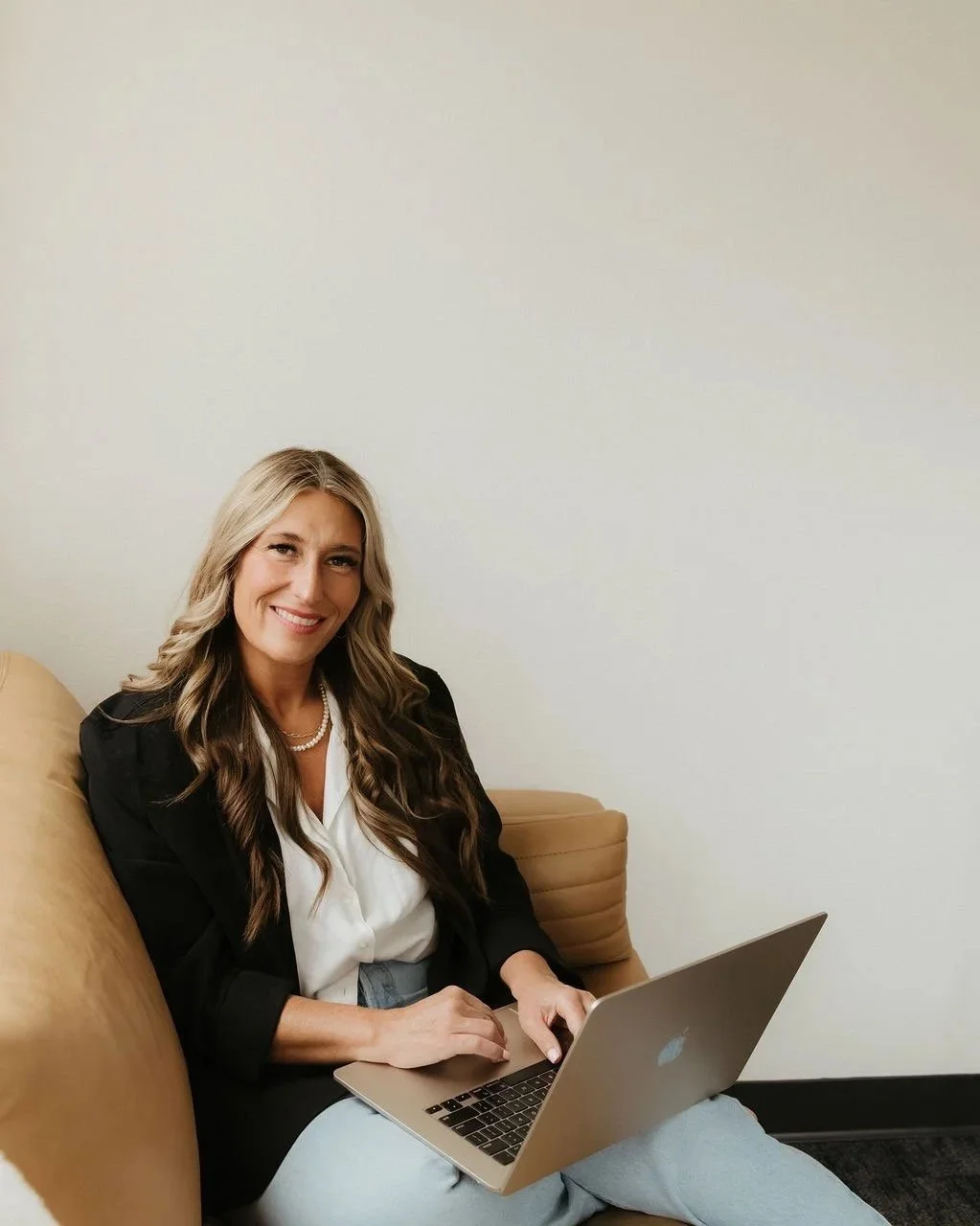 Woman sitting on a beige couch working on a MacBook laptop, smiling, against a plain wall.